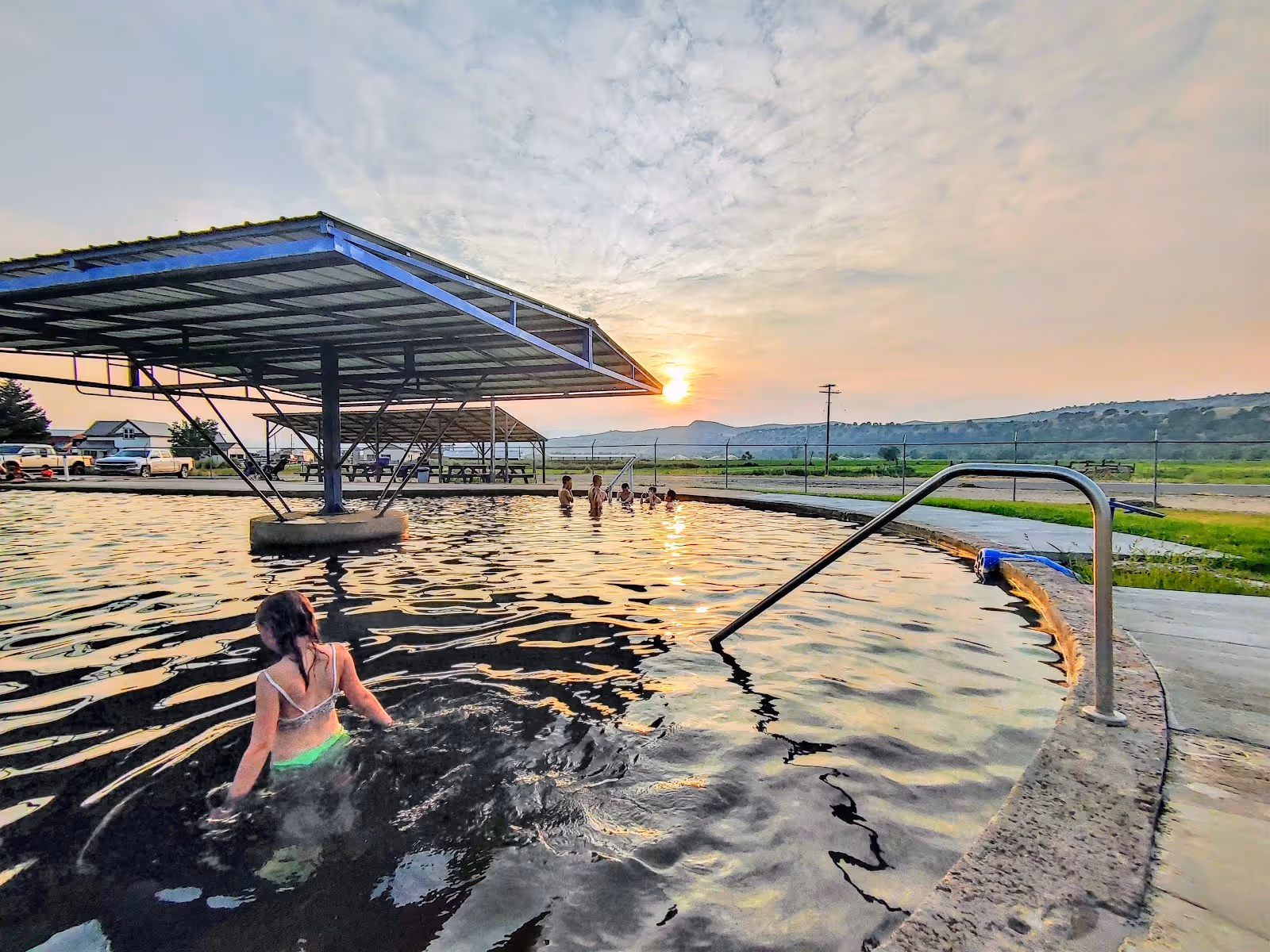 People bathing in the outdoor pool at Riverdale Resort Hot Springs at sunset near Preston, Idaho, with a metal railing and open landscape.