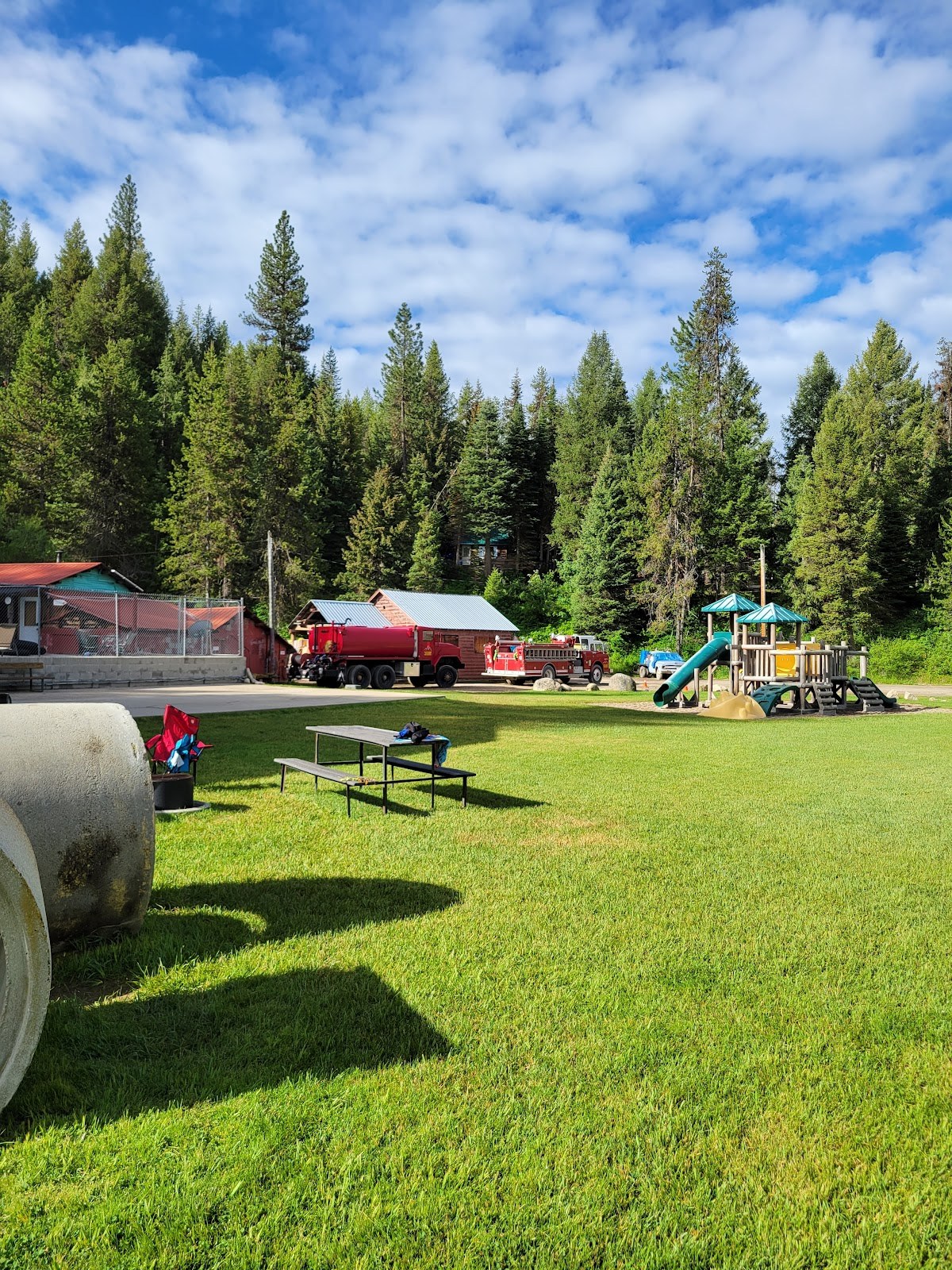 Green grassy picnic and playground area near Silver Creek Plunge in Crouch, Idaho, with fire trucks and forest in the background.