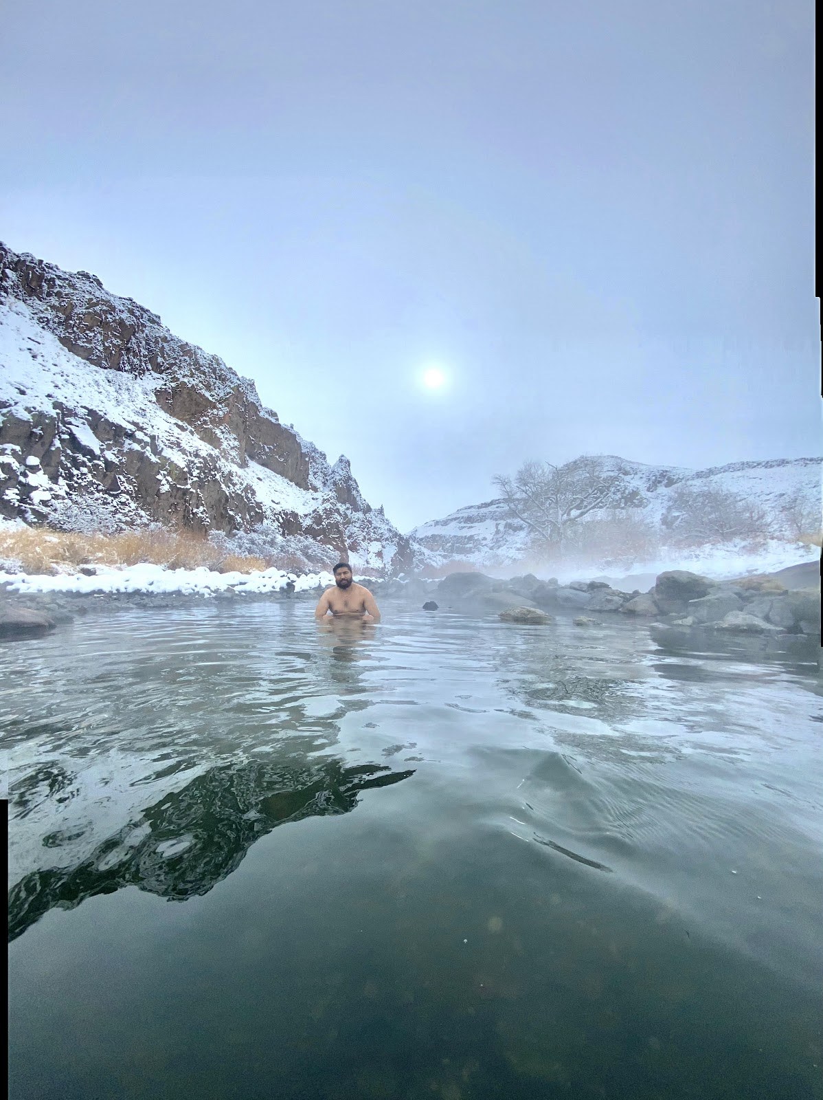 A person soaking in Snively Hot Springs surrounded by snow-covered rocky cliffs near Adrian, Idaho, United States with steam rising.