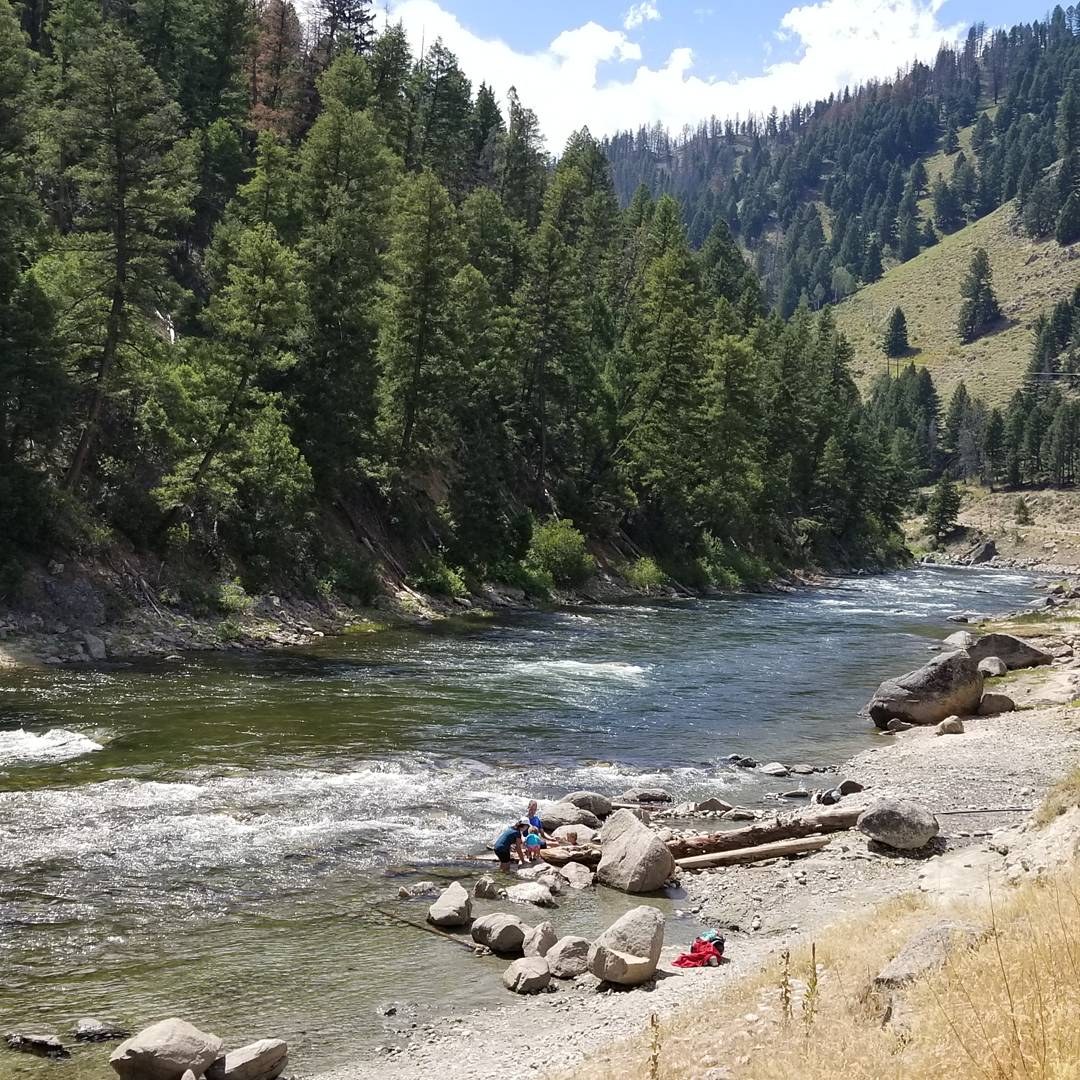 A natural hot spring area beside a flowing river with people enjoying the water and pine trees on hills at Sunbeam Hot Springs, Idaho.
