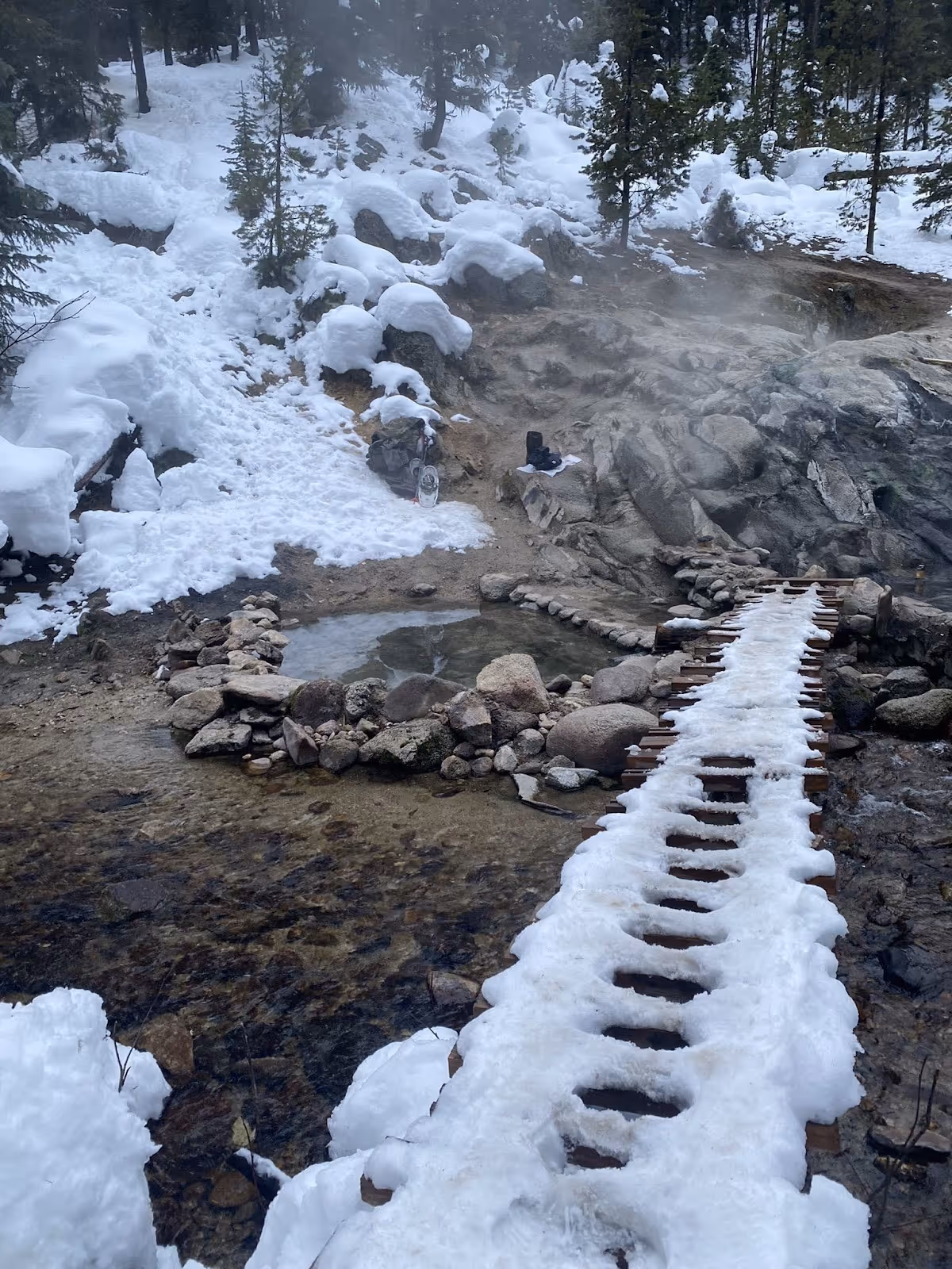 Snow-covered wooden boardwalk leading to steaming stone pools at Trail Creek Hot Springs amid snowy forest near Cascade, Idaho.
