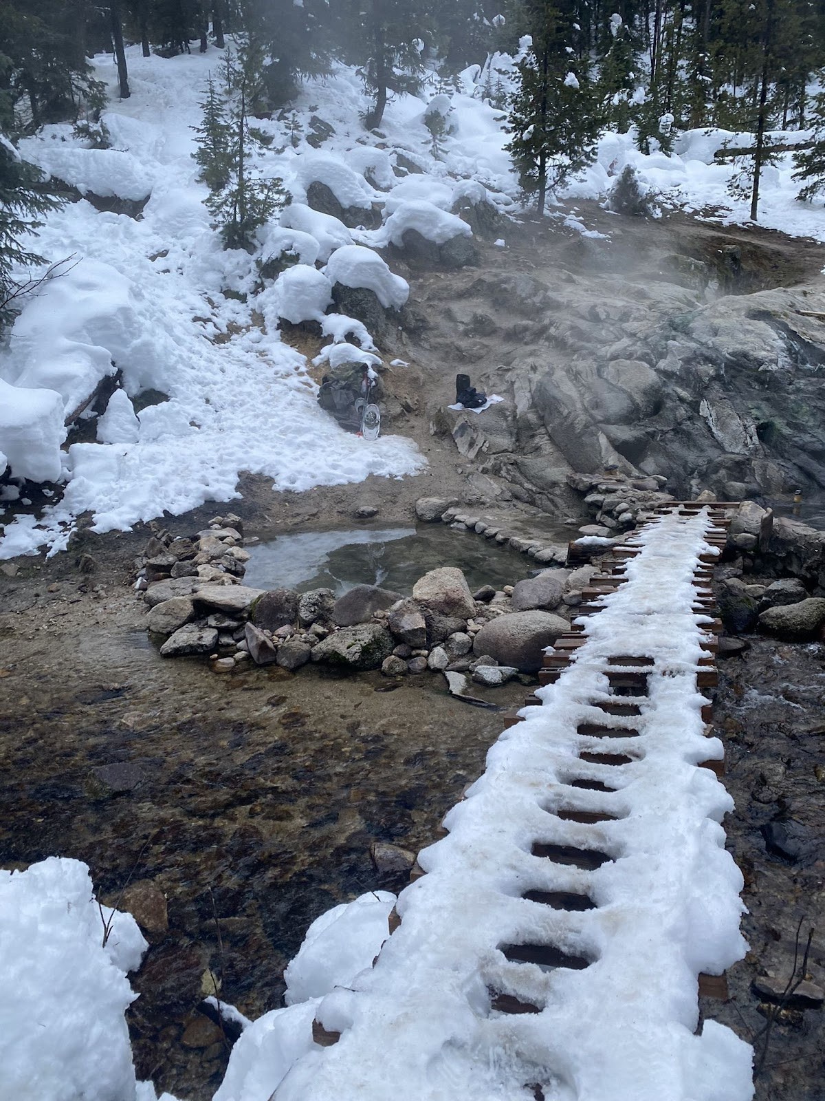 Snow-covered wooden boardwalk leading to steaming stone pools at Trail Creek Hot Springs amid snowy forest near Cascade, Idaho.