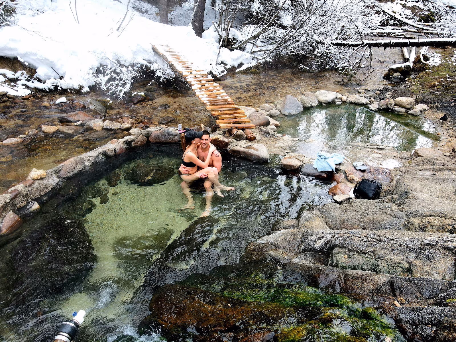Couple embracing in the warm stone pools of Trail Creek Hot Springs near Cascade, Idaho, surrounded by light snow and a narrow wood bridge.