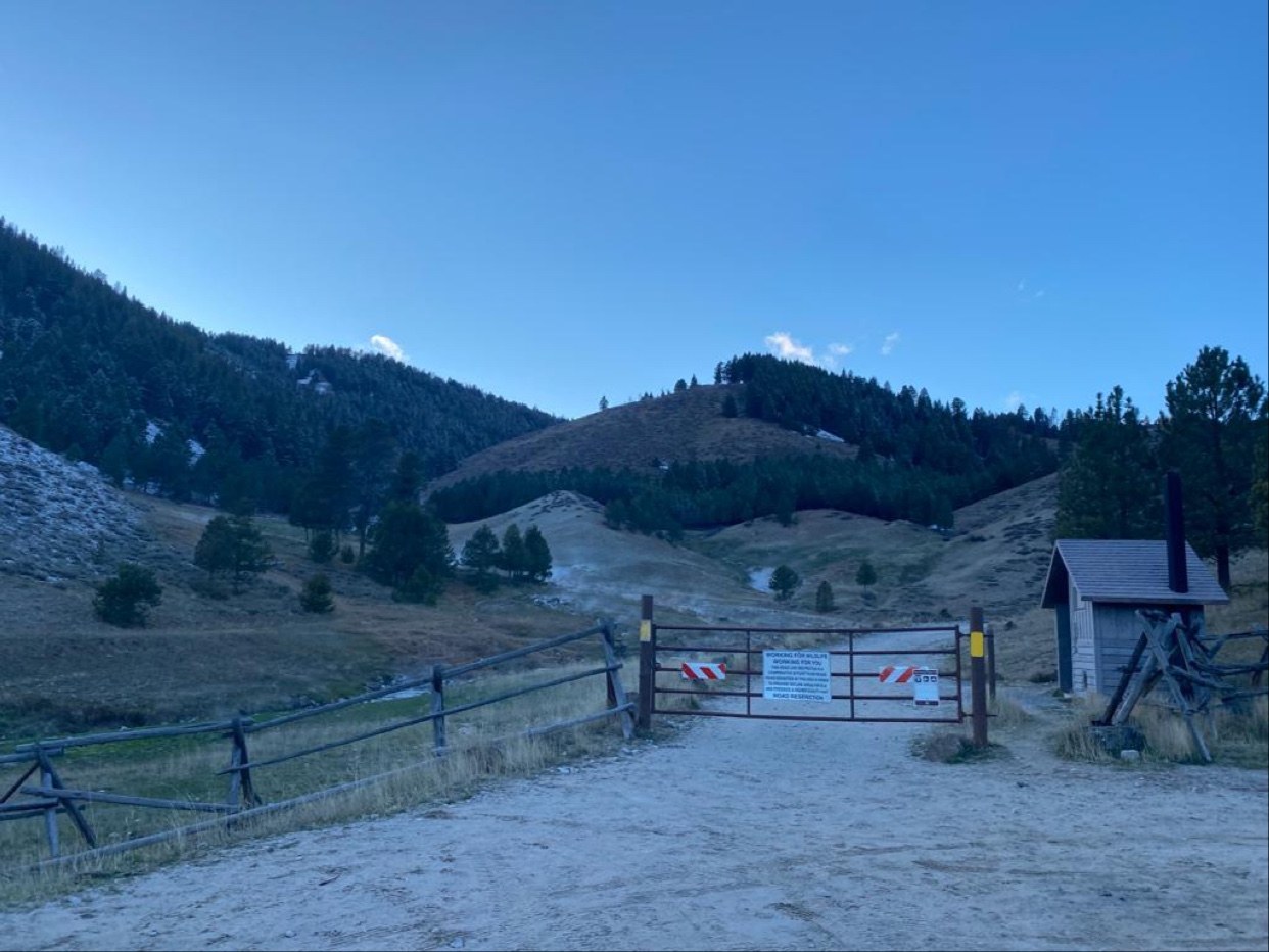 Entrance gate with informational signs to Worswick Hot Springs, set against forested hills under a clear blue sky near Fairfield, Idaho.