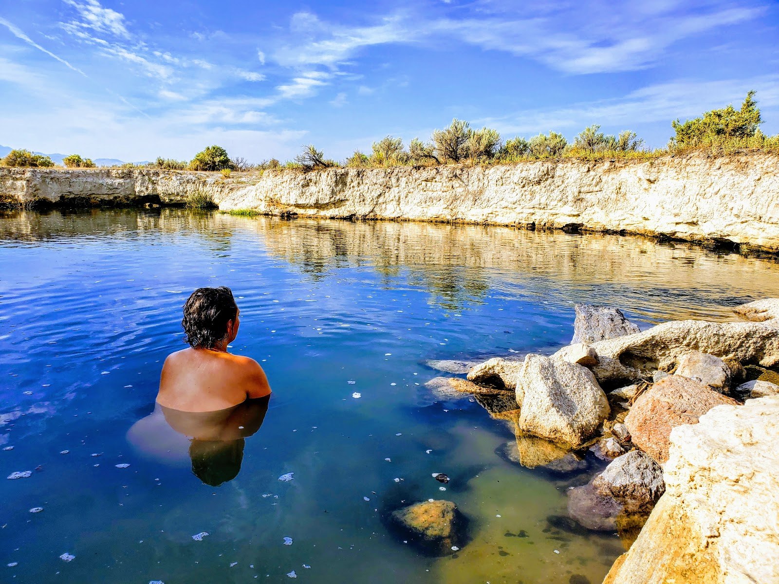 Person relaxing in Bog Hot Springs, an outdoor natural pool near Denio, Nevada, under a clear blue sky with rocky edges and shrubs