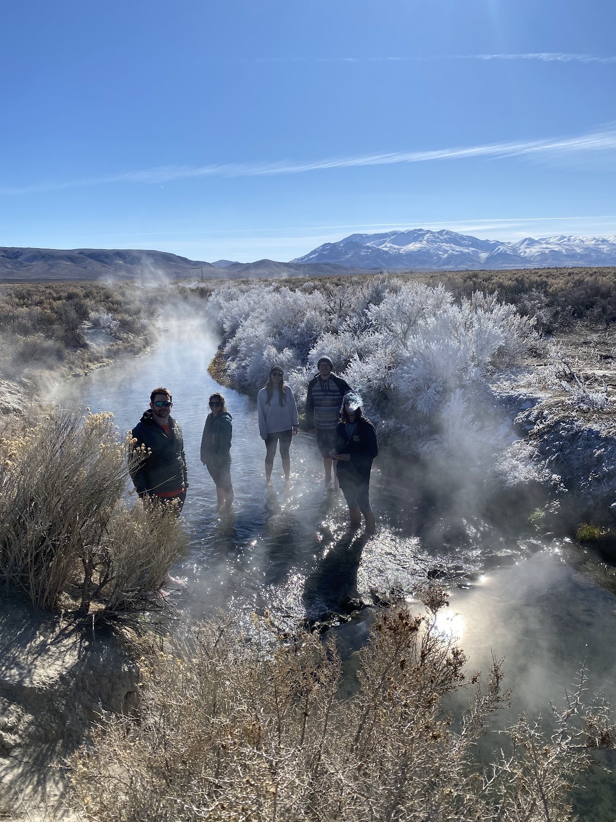 Group of people standing in a steaming creek at Bog Hot Springs with snowy shrubs and mountain views near Denio, Nevada