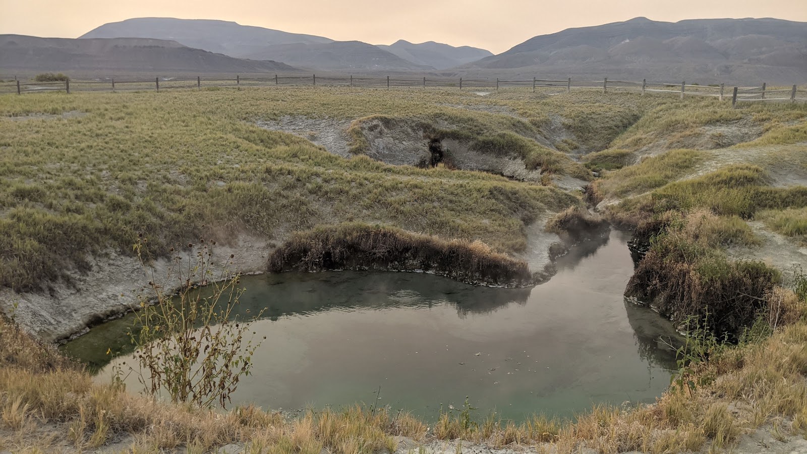 Steam gently rising from the calm waters of Double Hot Springs amid grassy terrain and fenced fields near Gerlach, Nevada