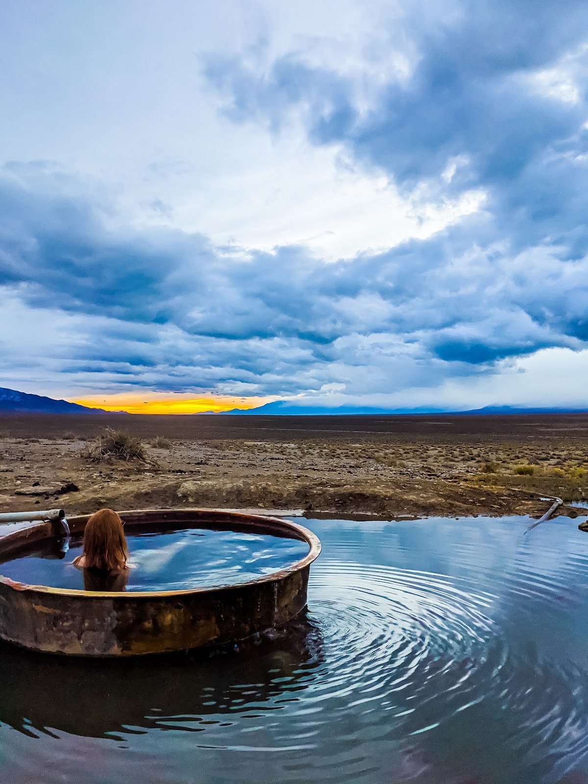 Person soaking in a round metal tub at Spencer Hot Springs with expansive desert and cloudy sunset near Austin, Nevada, United States.