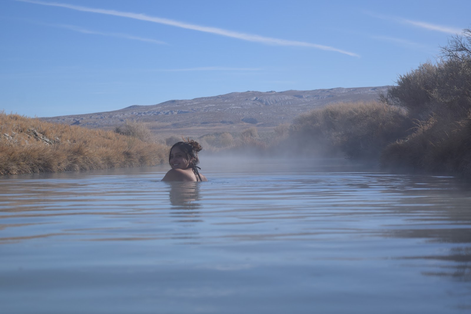 Person relaxing in steaming waters of Trego Hot Springs river with arid bushes and mountains near Gerlach, Nevada.