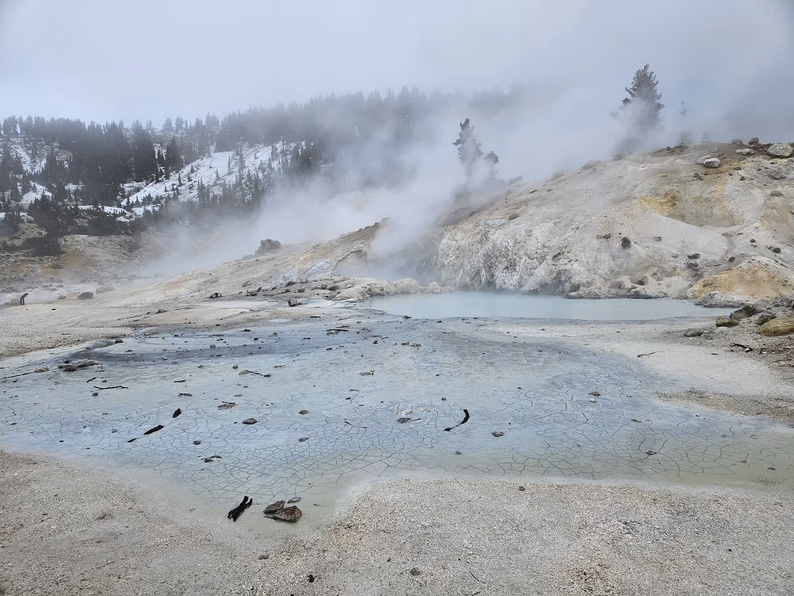 Foggy scene at Bumpass Hell Trail showing a cracked earth mineral pool with steam rising and snow dusting the nearby forested hills.