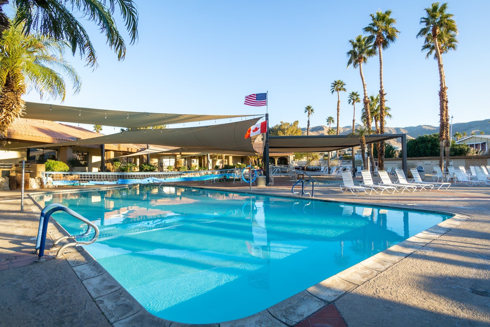 Wide rectangular outdoor pool at Caliente Springs Resort in Desert Hot Springs California with white lounge chairs and two flags on a pole.