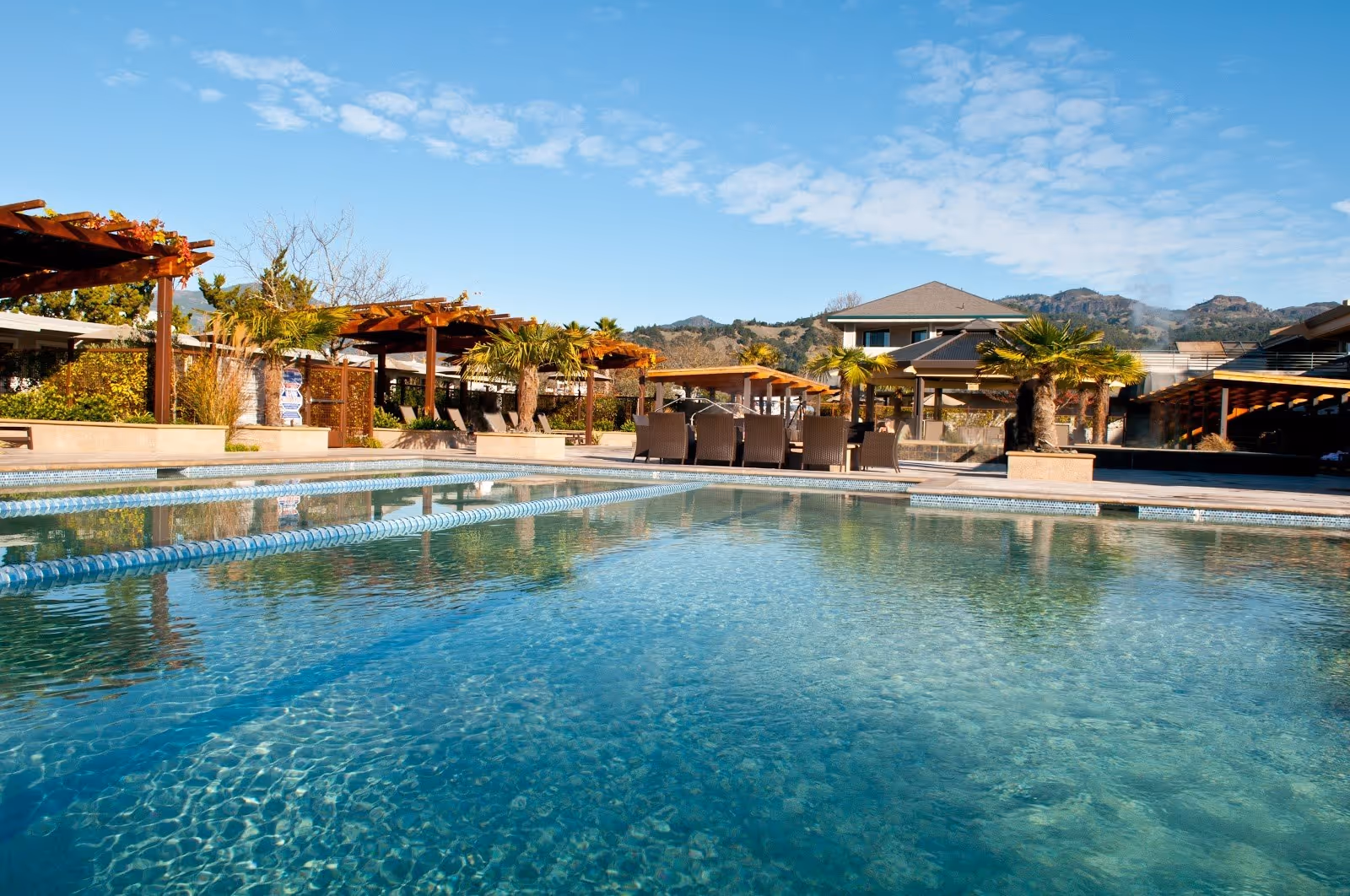 Clear blue outdoor mineral pool at Calistoga Spa Hot Springs with lounge chairs and wooden pergolas under a partly cloudy sky.