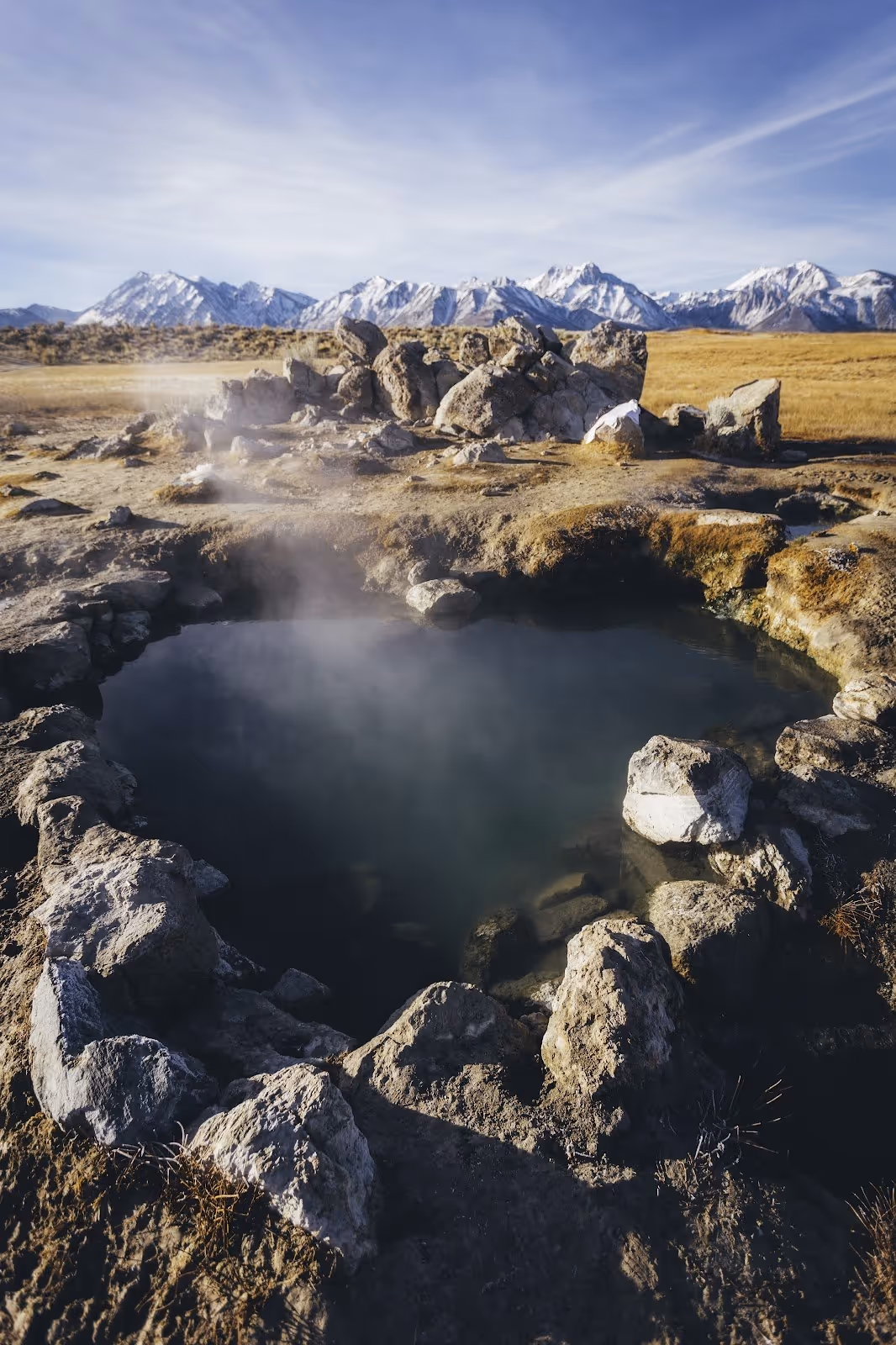 Steam rising from a rocky mineral pool of Crowley Wild Willy's Hot Spring with snow-capped mountains visible across the dry grassland near.