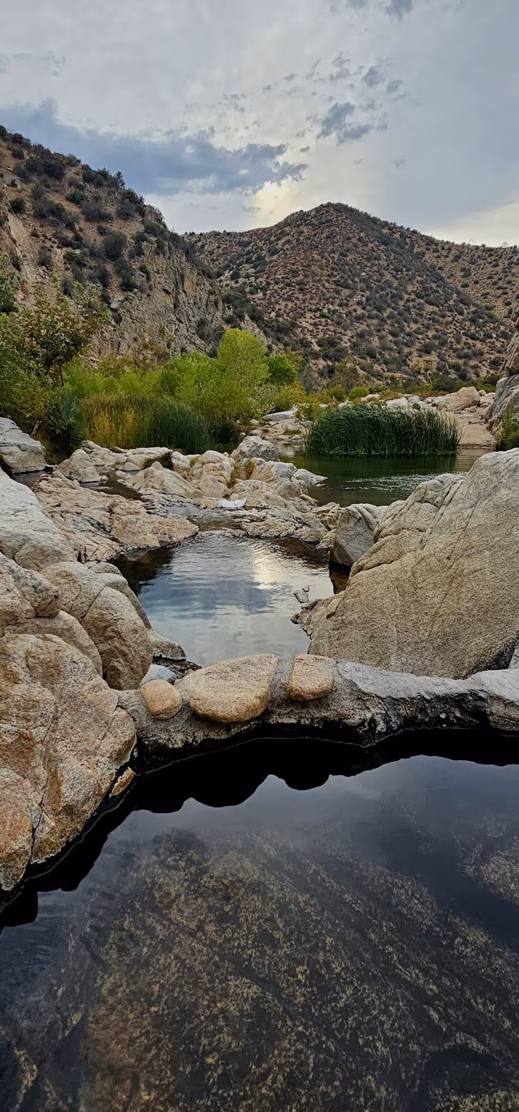 Natural hot spring pool surrounded by large rocks and green bushes with hills in the background at Deep Creek Hot Springs, California.