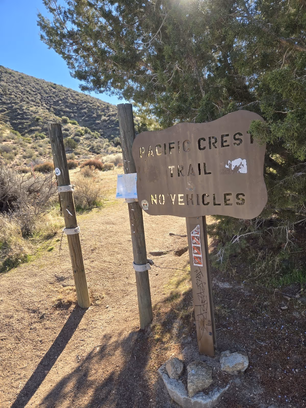 Weathered wooden sign for Pacific Crest Trail at a dirt path trailhead with hills and sparse vegetation near Deep Creek Hot Springs, Califo.