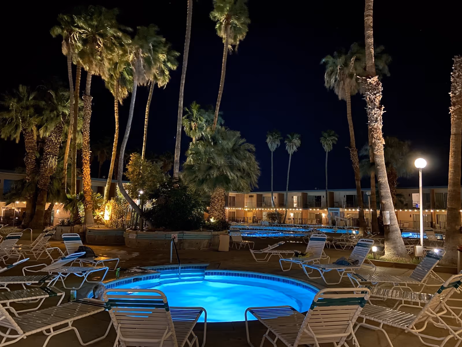 Night view of illuminated blue hot spring soaking pool at Desert Hot Springs Spa Hotel with empty lounge chairs and palm trees under dark s.