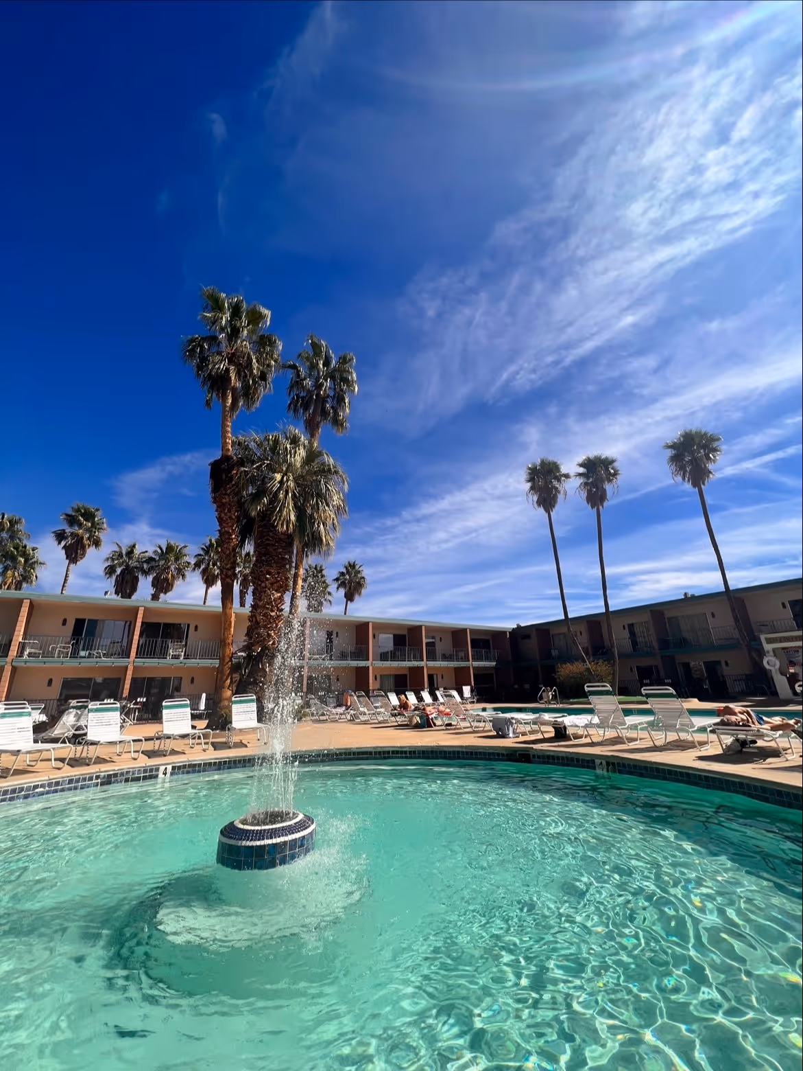 Fountain sprays water in the center of a turquoise-tiled outdoor spa pool with sunlit palm trees and hotel balconies in Desert Hot Springs.