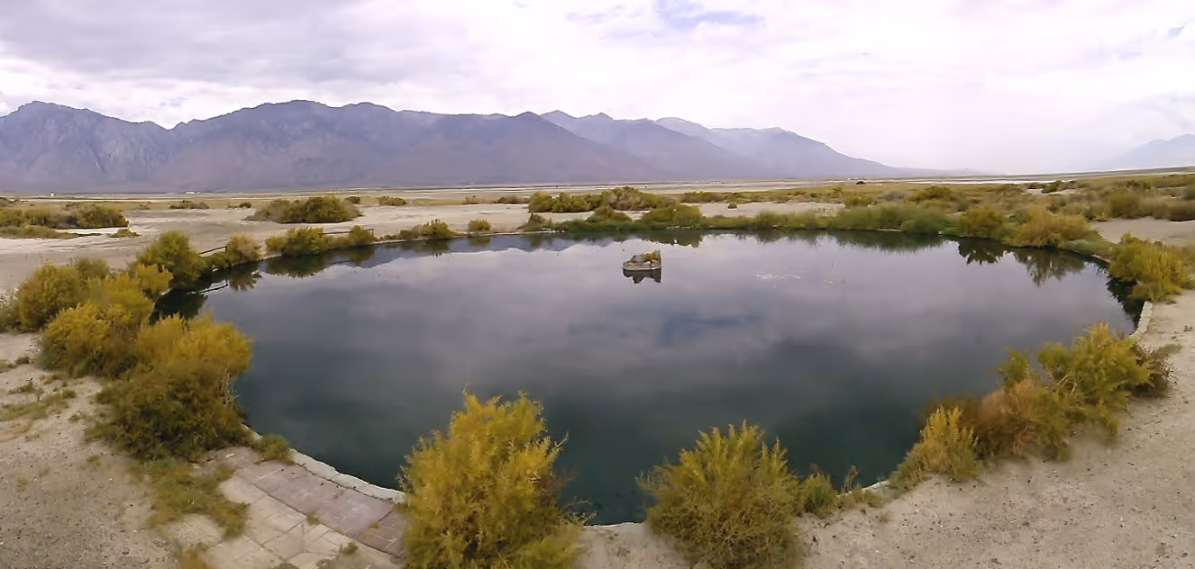 The calm water of Dirty Sock Hot Springs reflects cloudy skies and yellow-green shrubs with distant mountain ranges on the horizon close to.