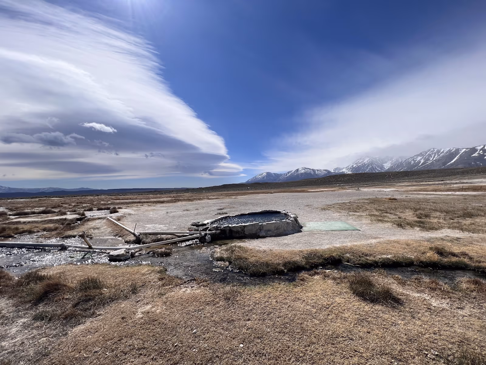 Stone-edged hot spring pool at Hilltop Pulky's Pool with dry grassy field and snow-capped mountains under a blue sky in California.