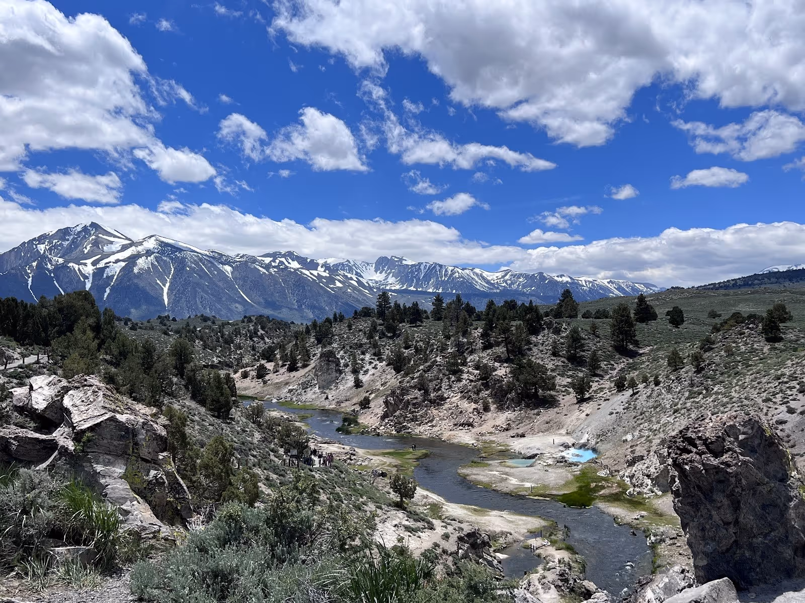 Wide view of Hot Creek Geologic Site valley with snow capped mountains in the distance and scattered trees on hillsides.