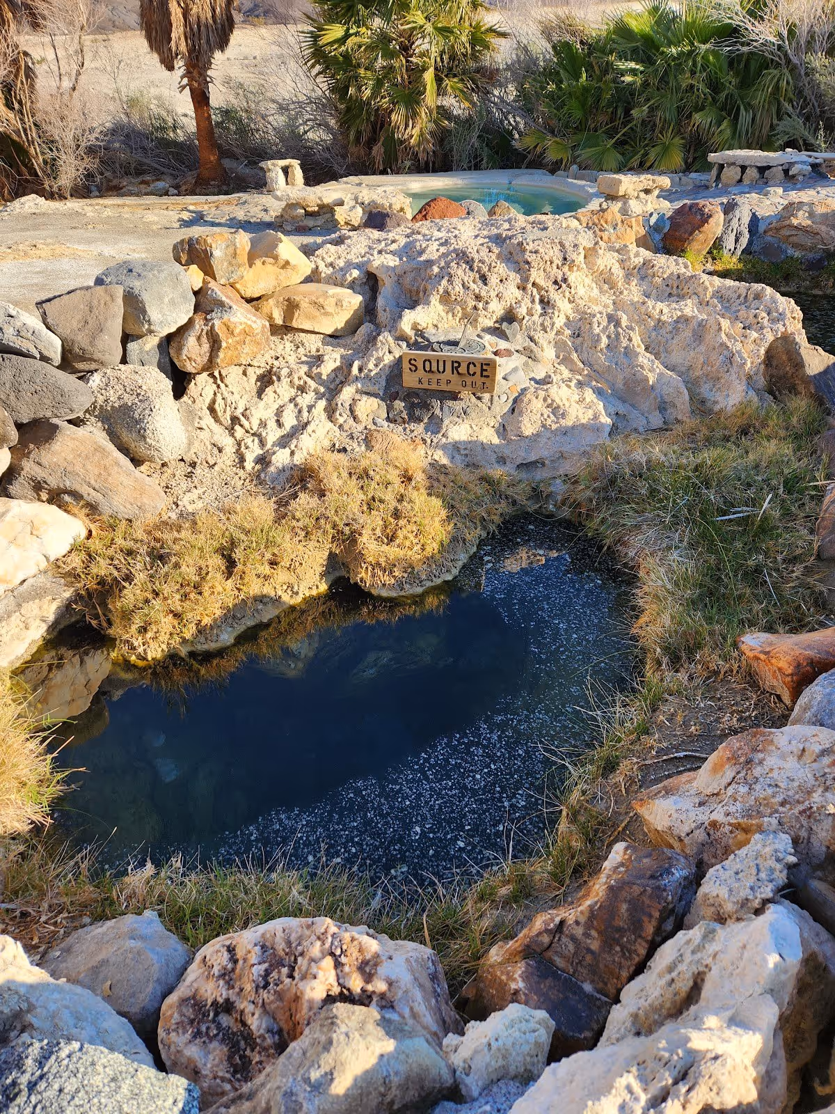 Rock-lined natural hot spring pool labeled source with a keep out sign surrounded by dry grass and desert plants near Big Pine california.