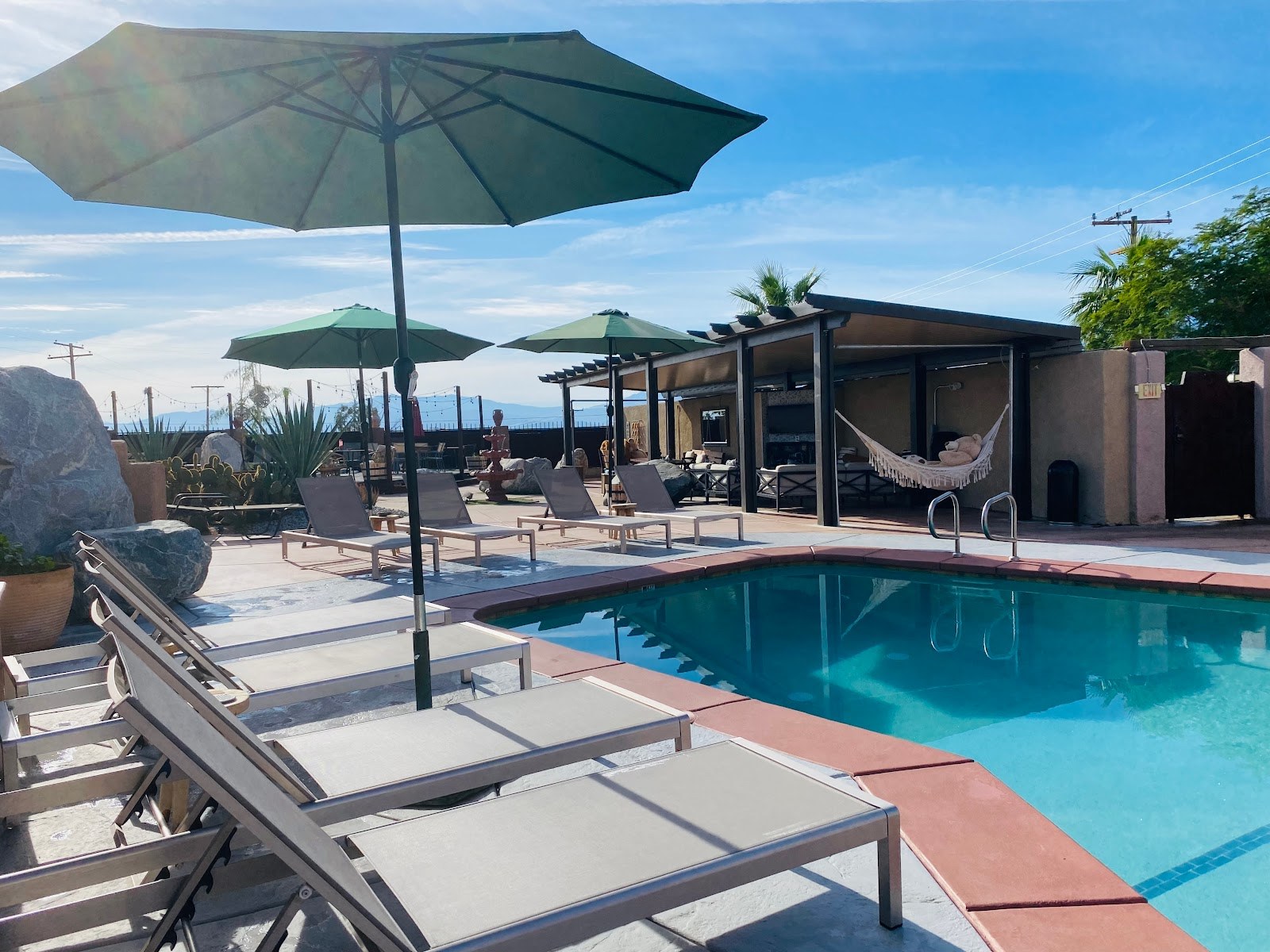 Bright blue soaking pool at Mi Kasa Hot Springs with multiple green sun umbrellas and beige lounge chairs on the concrete deck under a sunn.