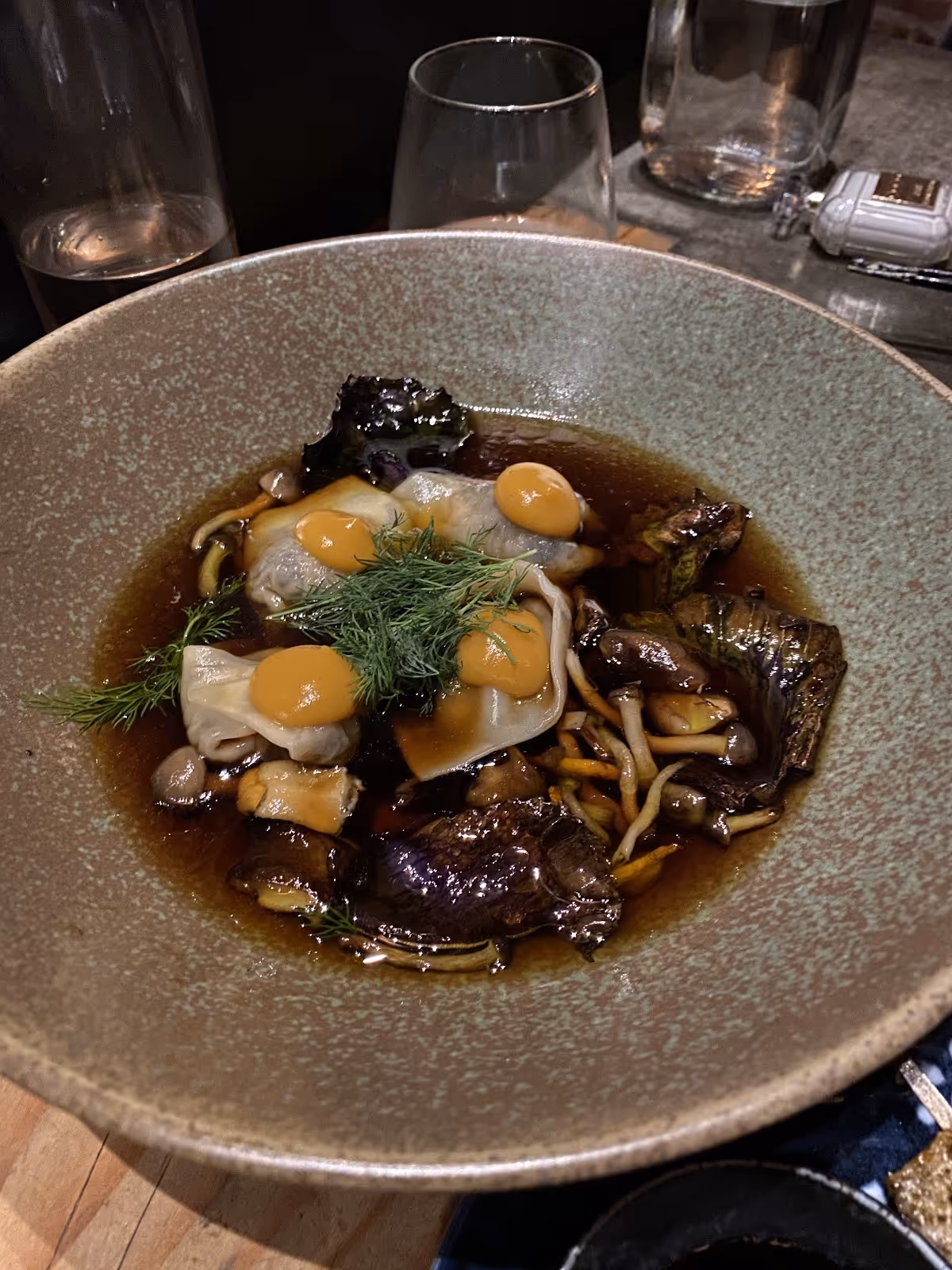 A dark gray bowl with mushroom and herb garnished dish on a wooden table next to empty glasses and a bottle.