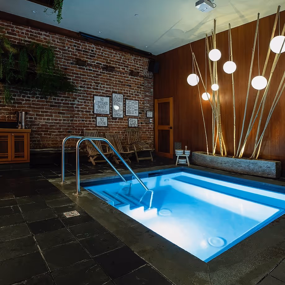 Lit indoor soaking pool at Onsen SF in San Francisco showing metal steps, brick wall, wood seats, and modern hanging lights.