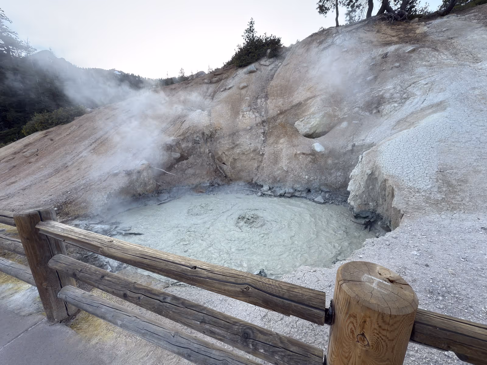 Milky hot spring mud pool encircled by a wooden fence with steam and rocky hillsides at Sulphur Works, California.
