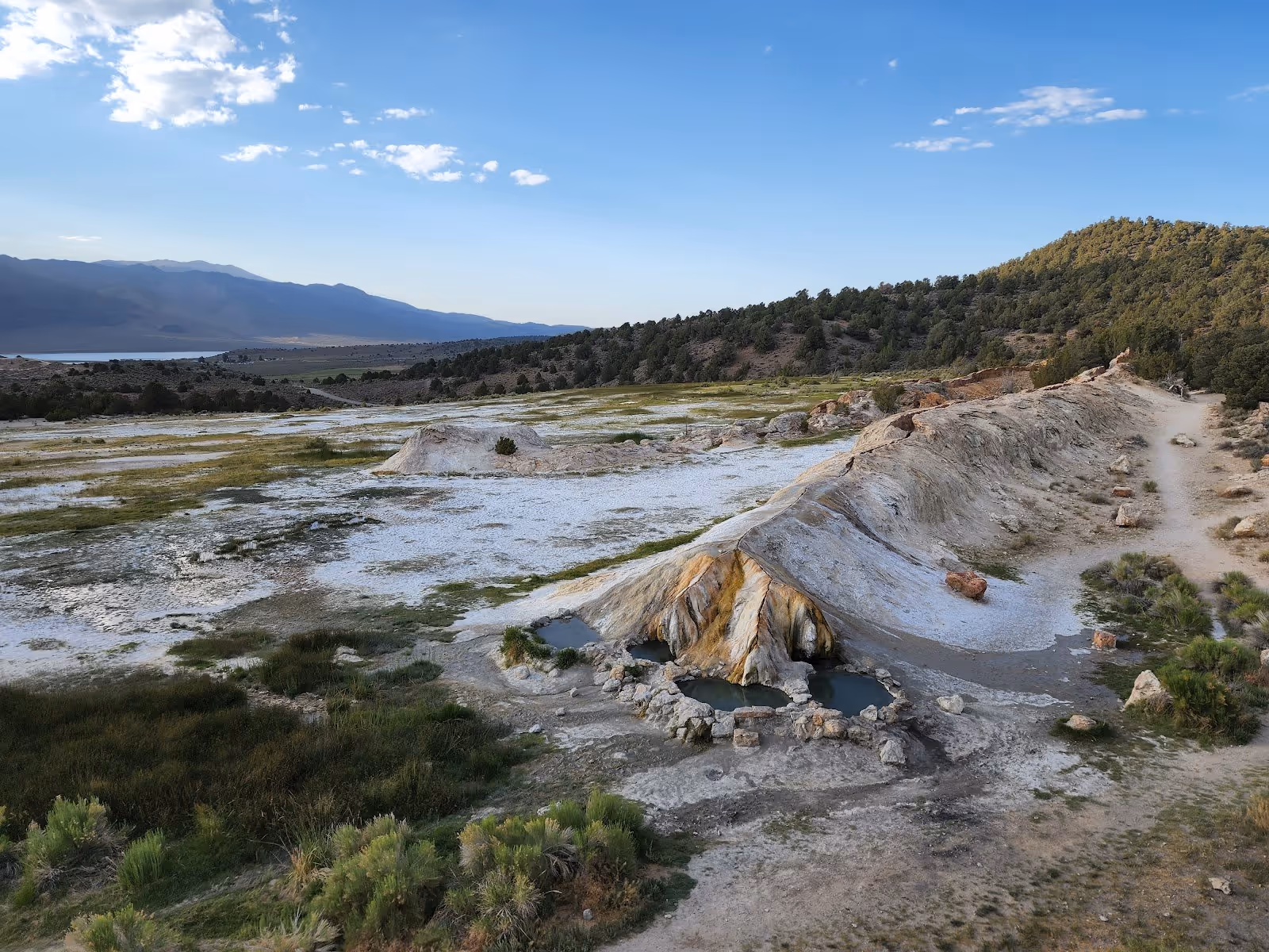 Wide view of Travertine hot springs showing multiple mineral pools surrounded by grassy and rocky terrain with mountains in the background.