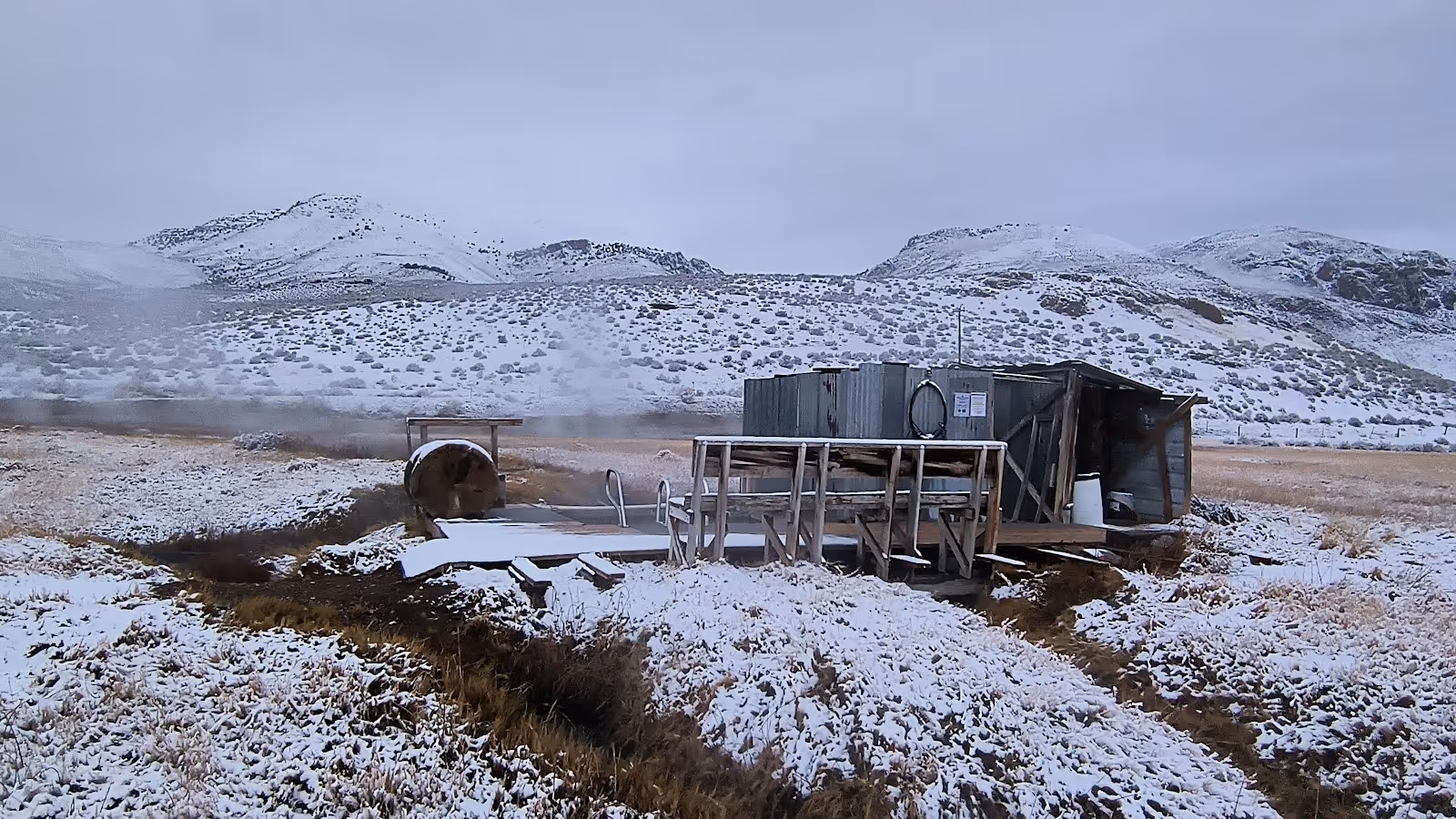 Snow-covered Alvord Hot Springs bathhouse and pool with visible steam, surrounded by snowy hills and white winter landscape near Princeton.