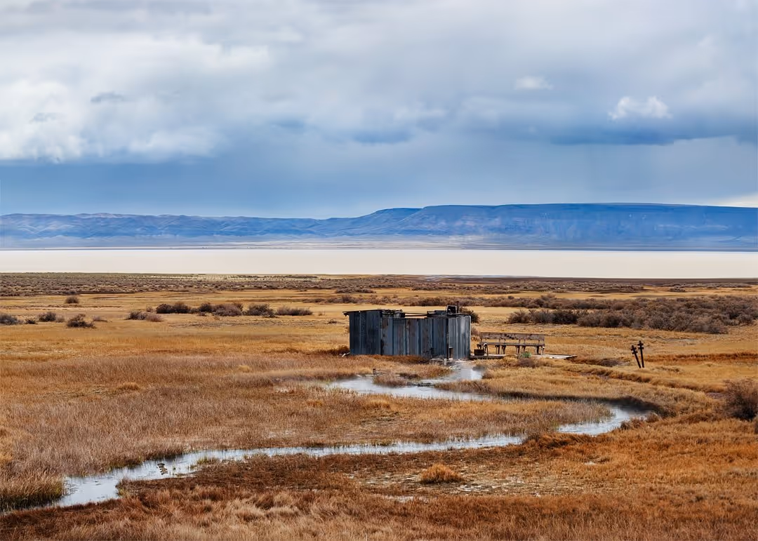 Rustic bathhouse by steaming hot spring on grassy prairie at Alvord Hot Springs, with distant mesa hills and cloudy sky in southeastern Ore.
