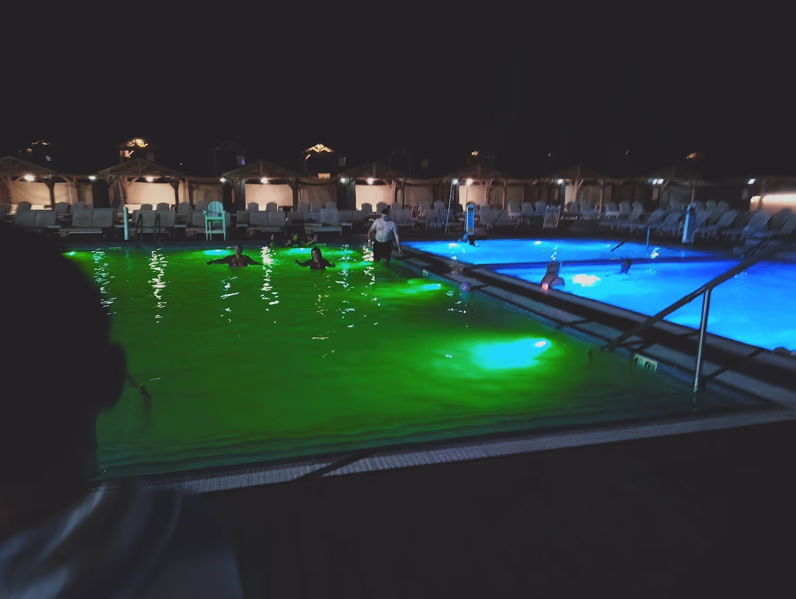 Night view of Kah-Nee-Ta Hot Springs soaking pools with glowing green and blue underwater lights and several bathers under soft ambient lig.