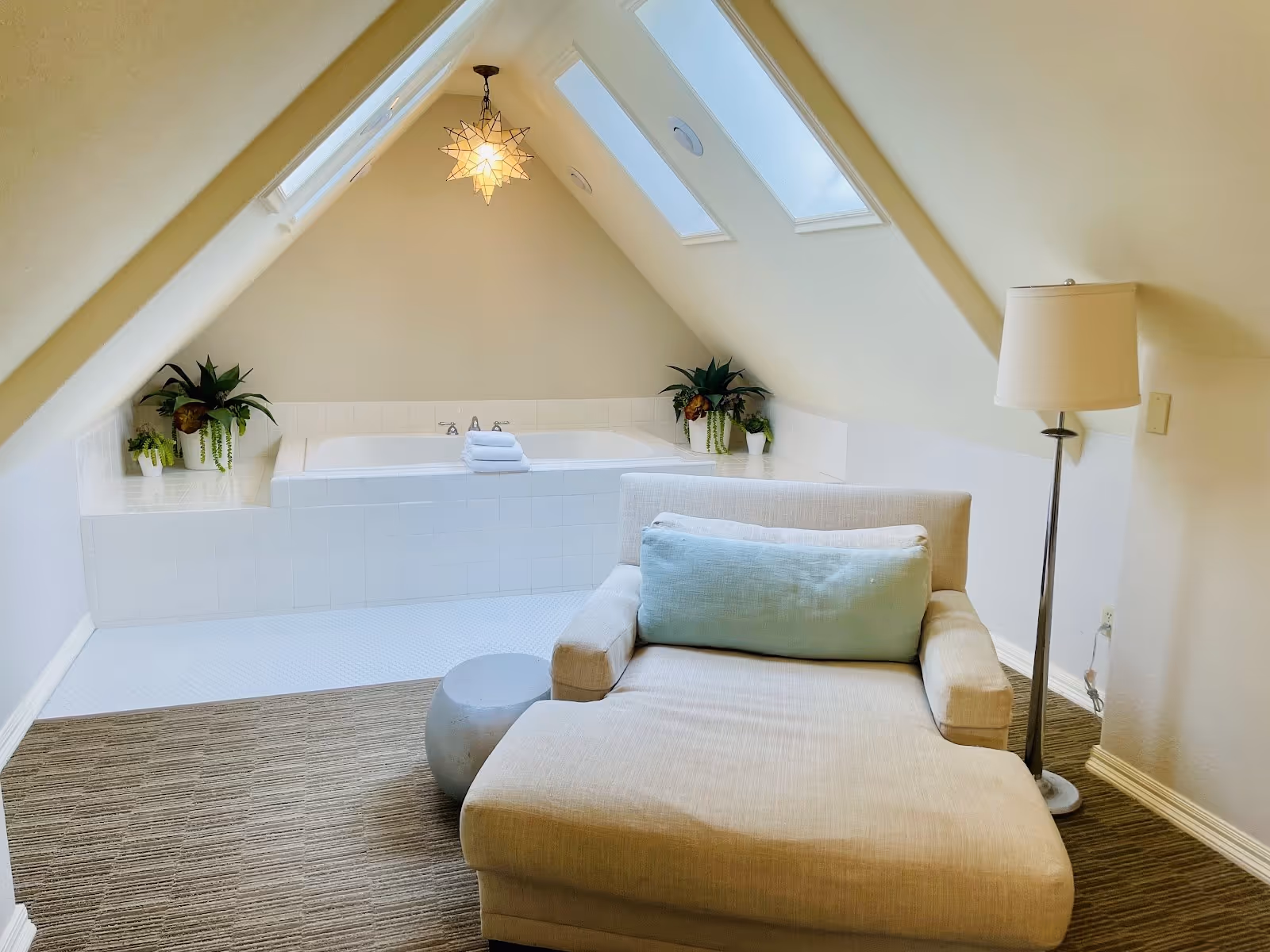 Bright indoor soaking tub under skylights with beige armchair, floor lamp, and plants in a cozy attic room.