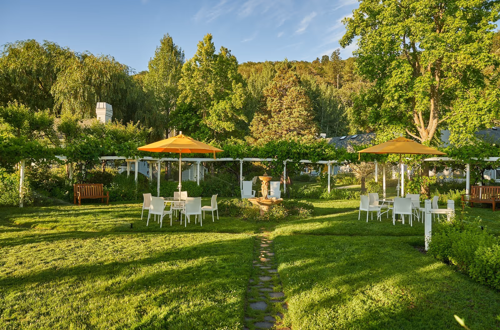 Green lawn area with white tables, yellow umbrellas, wooden benches and lush trees in the background on a sunny day.