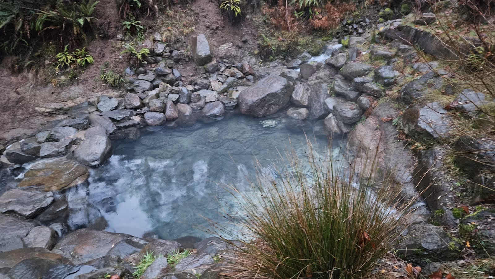 Clear mineral hot spring pool lined with rocks and plants reflecting overcast sky at Terwilliger Hot Springs near Blue River Oregon.