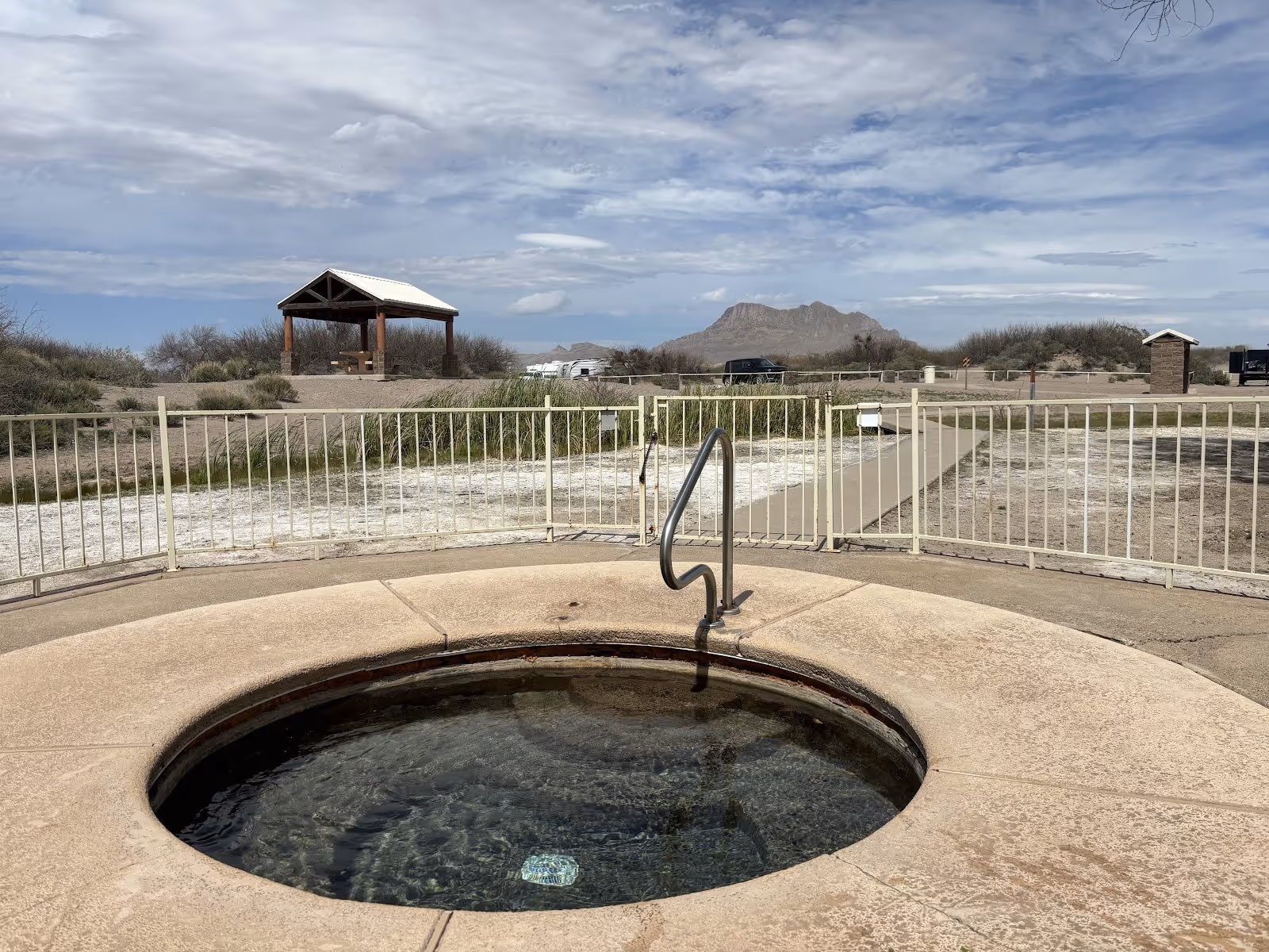 Round hot spring pool at Hot Well Dunes with metal handrail and fenced area, picnic pavilion and desert landscape beyond in Arizona.