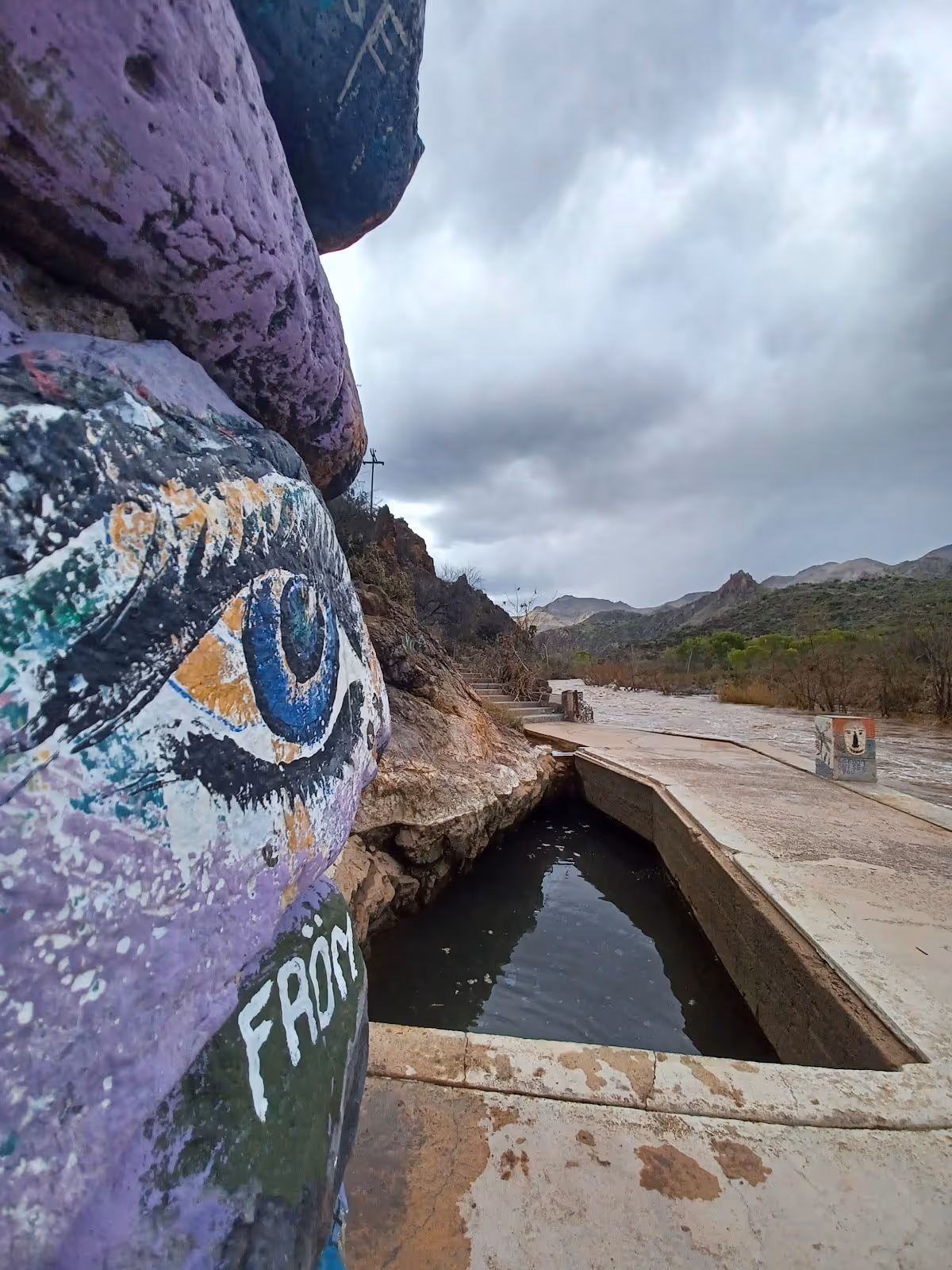 Close-up of painted stones with an eye design beside a rectangular hot spring pool and river under cloudy skies at Verde Hot Springs.