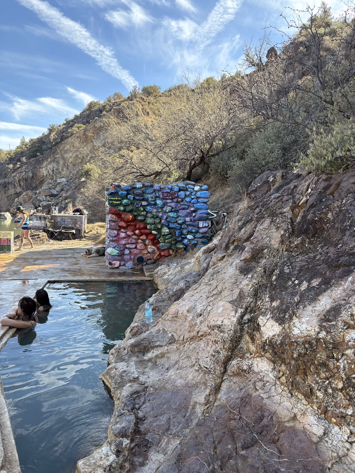 Several people relaxing in an outdoor hot spring pool at Verde Hot Springs, Arizona surrounded by rocky hills and painted rocks.