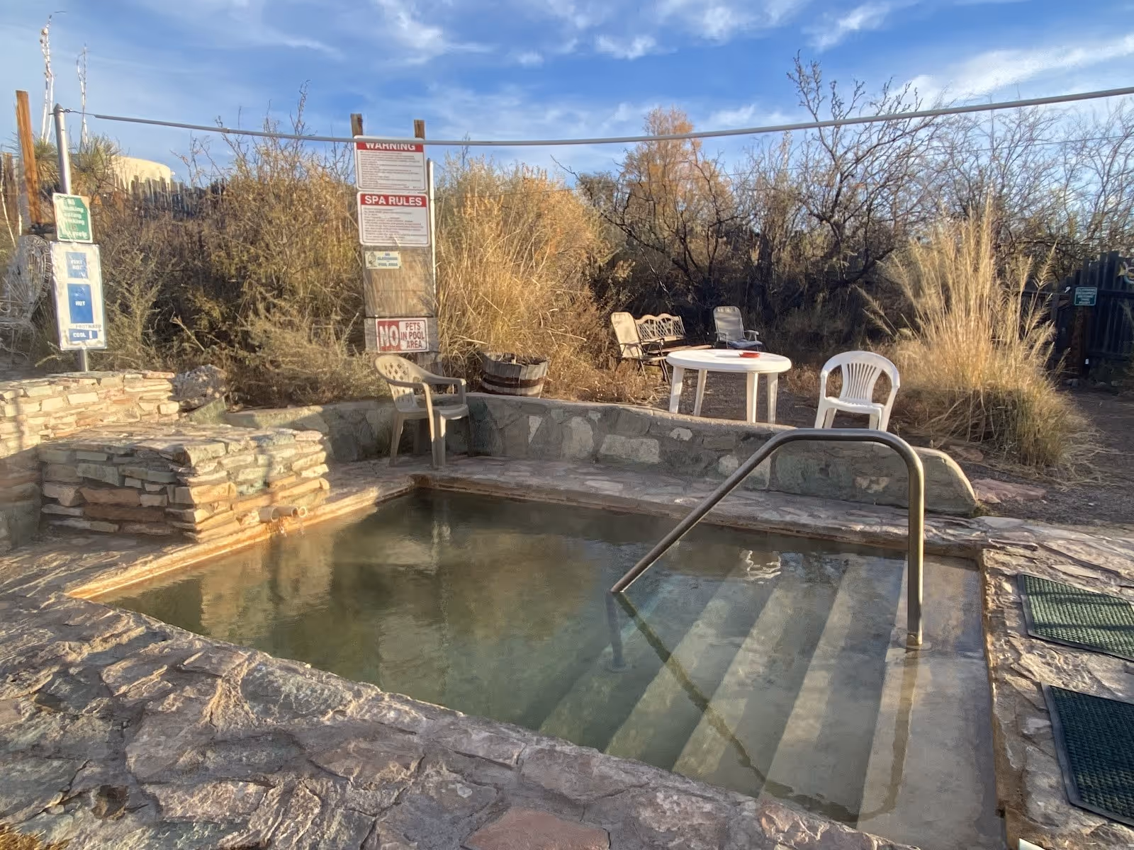 Small stone-bordered thermal pool at Faywood Hot Springs surrounded by dry brush and plastic chairs near Deming New Mexico.