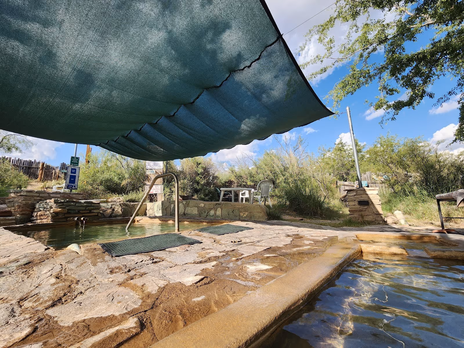 Two heated mineral pools at Faywood Hot Springs under a large shade cloth with a stone walkway and outdoor seating in New Mexico.