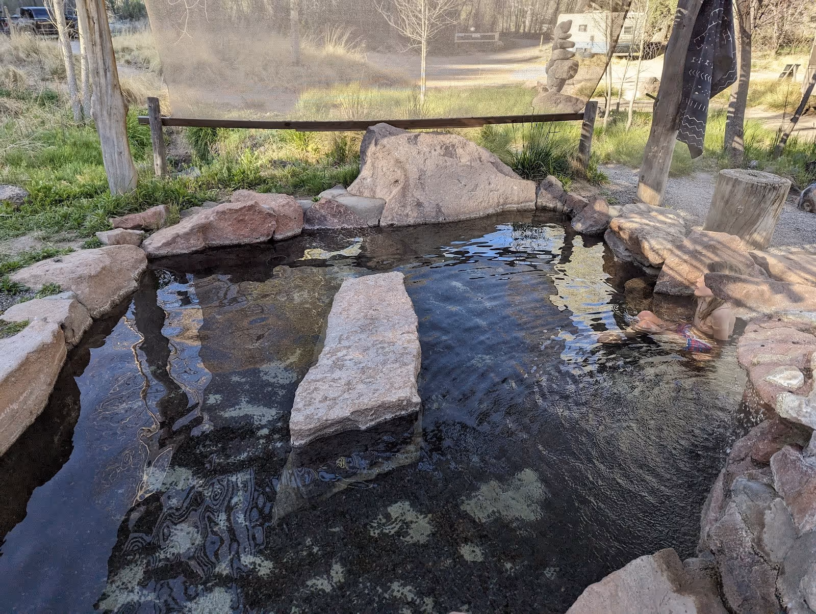 People soaking in a natural stone hot spring pool surrounded by grass and wooden posts at Gila Hot Springs near Silver City New Mexico.