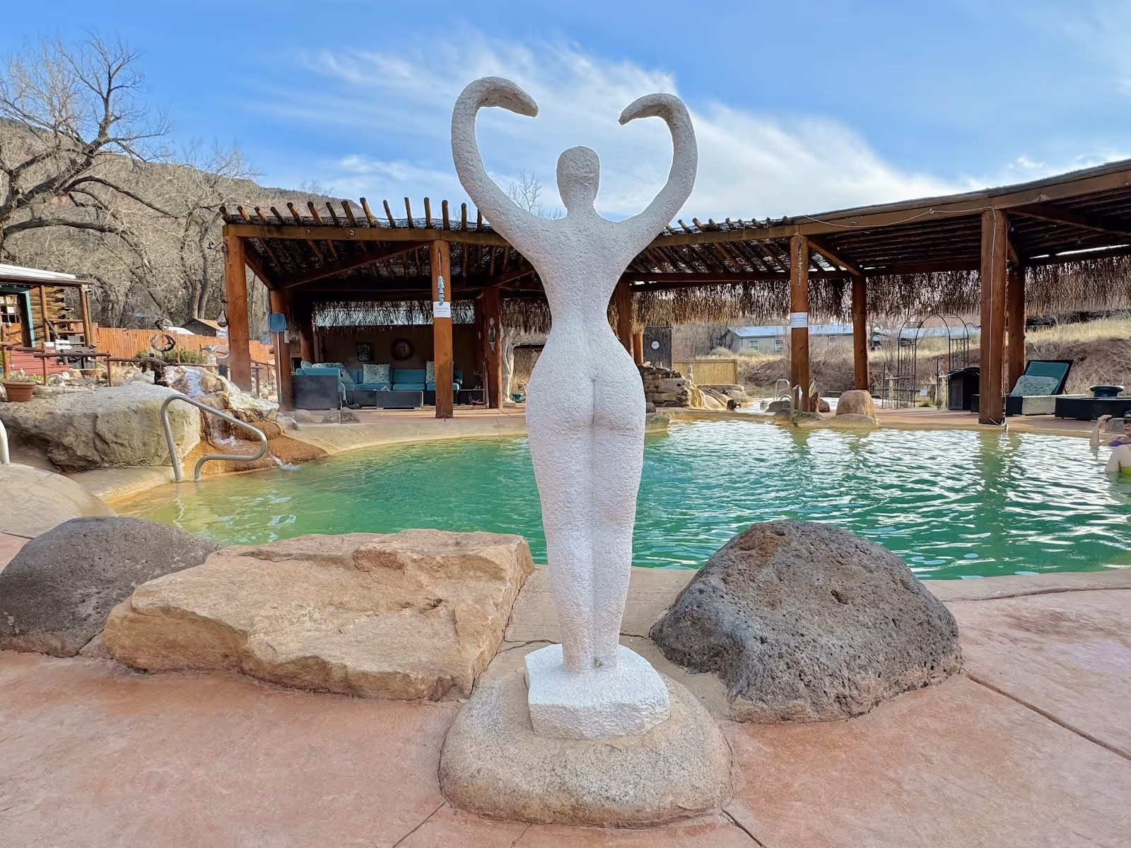 Jemez Hot Springs pool with turquoise water features a white sculpture of a figure raising arms near stone seating and covered lounging are.