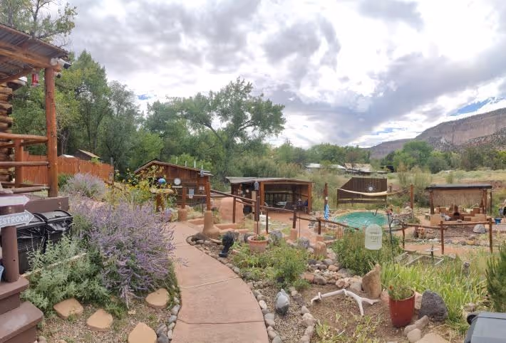 Panoramic view of Jemez Hot Springs soaking area with a garden pathway, several small wooden shelters, and lush trees in Jemez Springs New.