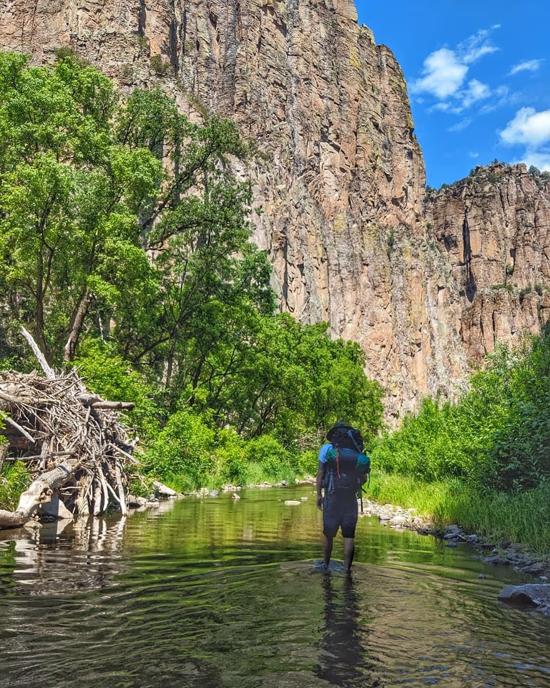 A person with a large backpack wades through a shallow stream with tall cliffs and green trees in the background under a blue sky.