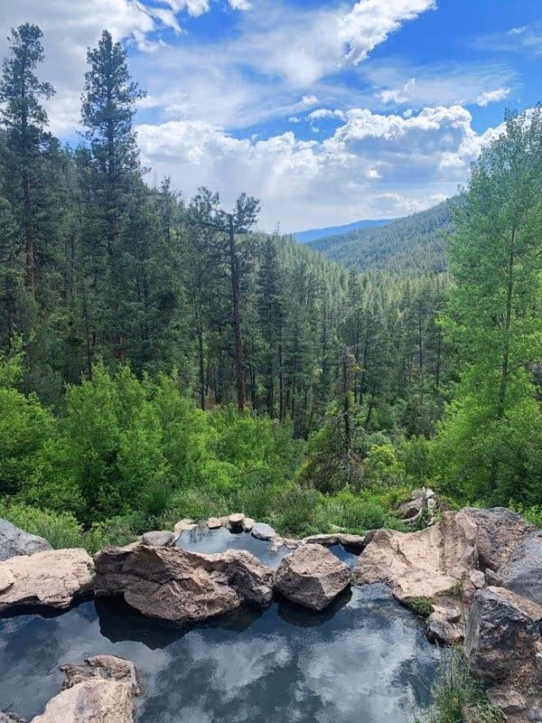 A natural rock-bordered hot spring pool reflecting a cloudy sky with surrounding forest and distant mountains at McCauley Warm Springs near.