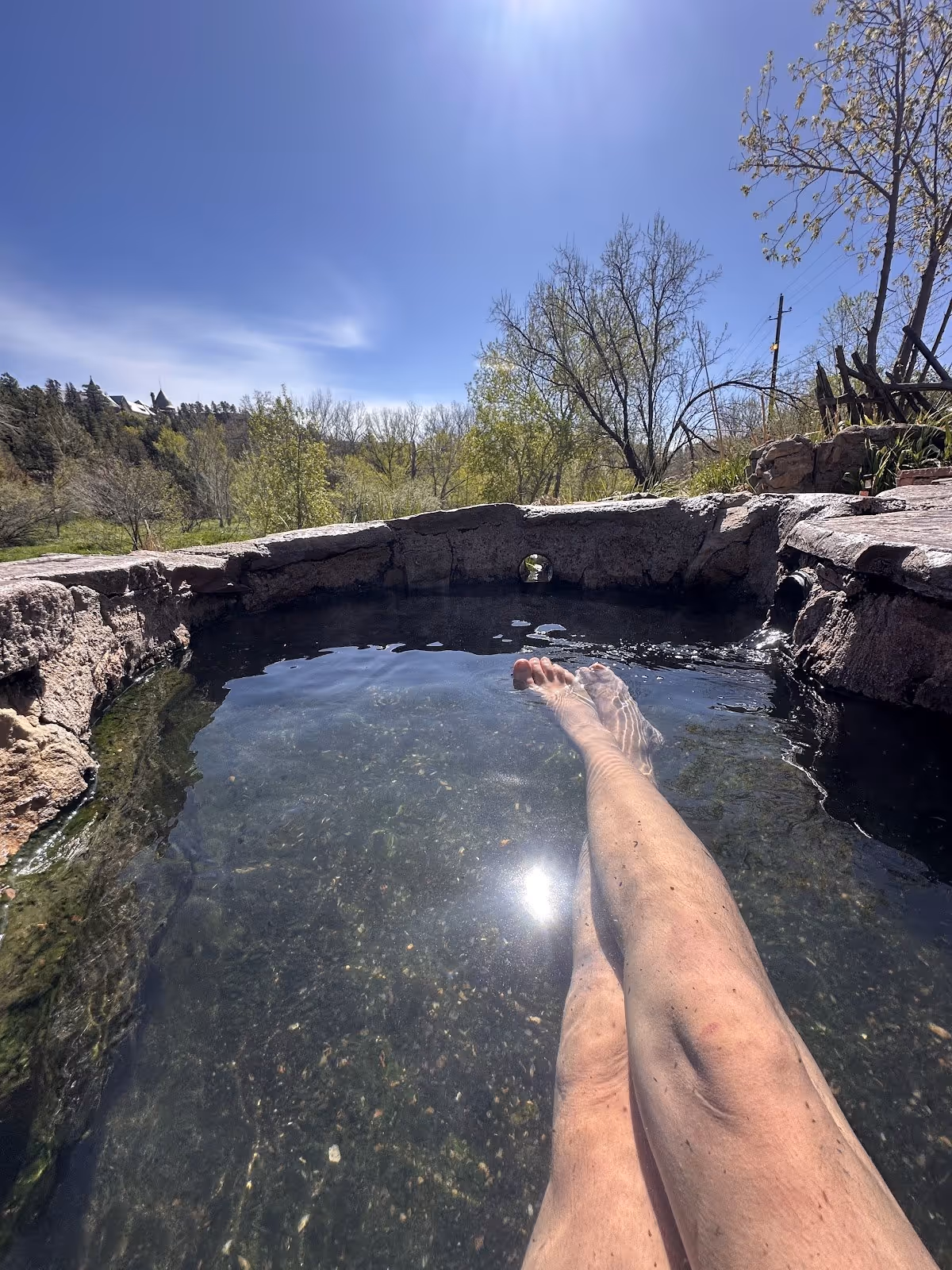 Legs submerged in a shallow round stone soaking pool at Montezuma Hot Springs with trees and blue sky visible beyond the wall.