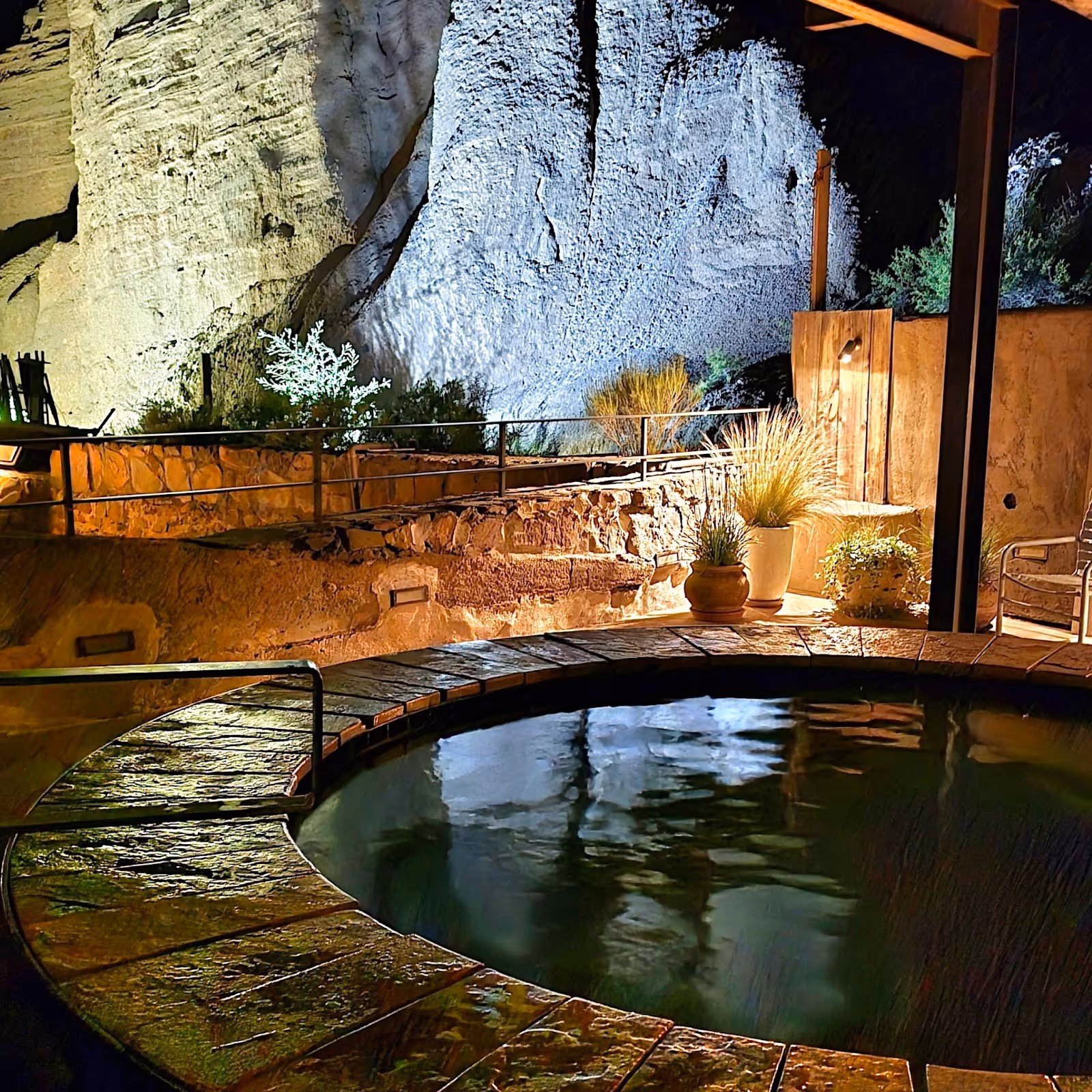 Round nighttime mineral pool at Ojo Caliente Mineral Springs with stone seating, greenery, and a rock wall lit by warm lights in New Mexico.