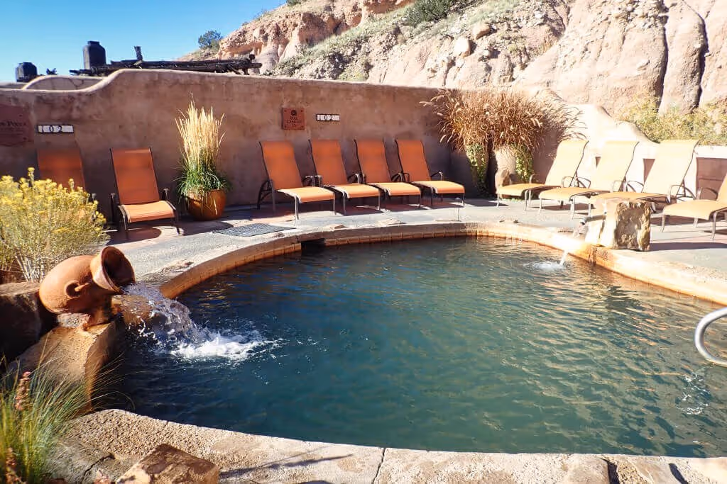 Outdoor hot spring pool at Ojo Caliente Mineral Springs with water flowing from a ceramic jug and lounge chairs in Espanola, New Mexico.