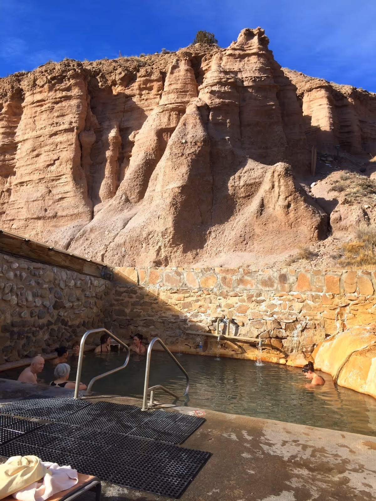 Soaking pool at Ojo Caliente Mineral Springs with stone walls and several people relaxing beside tall rocky cliffs in New Mexico.